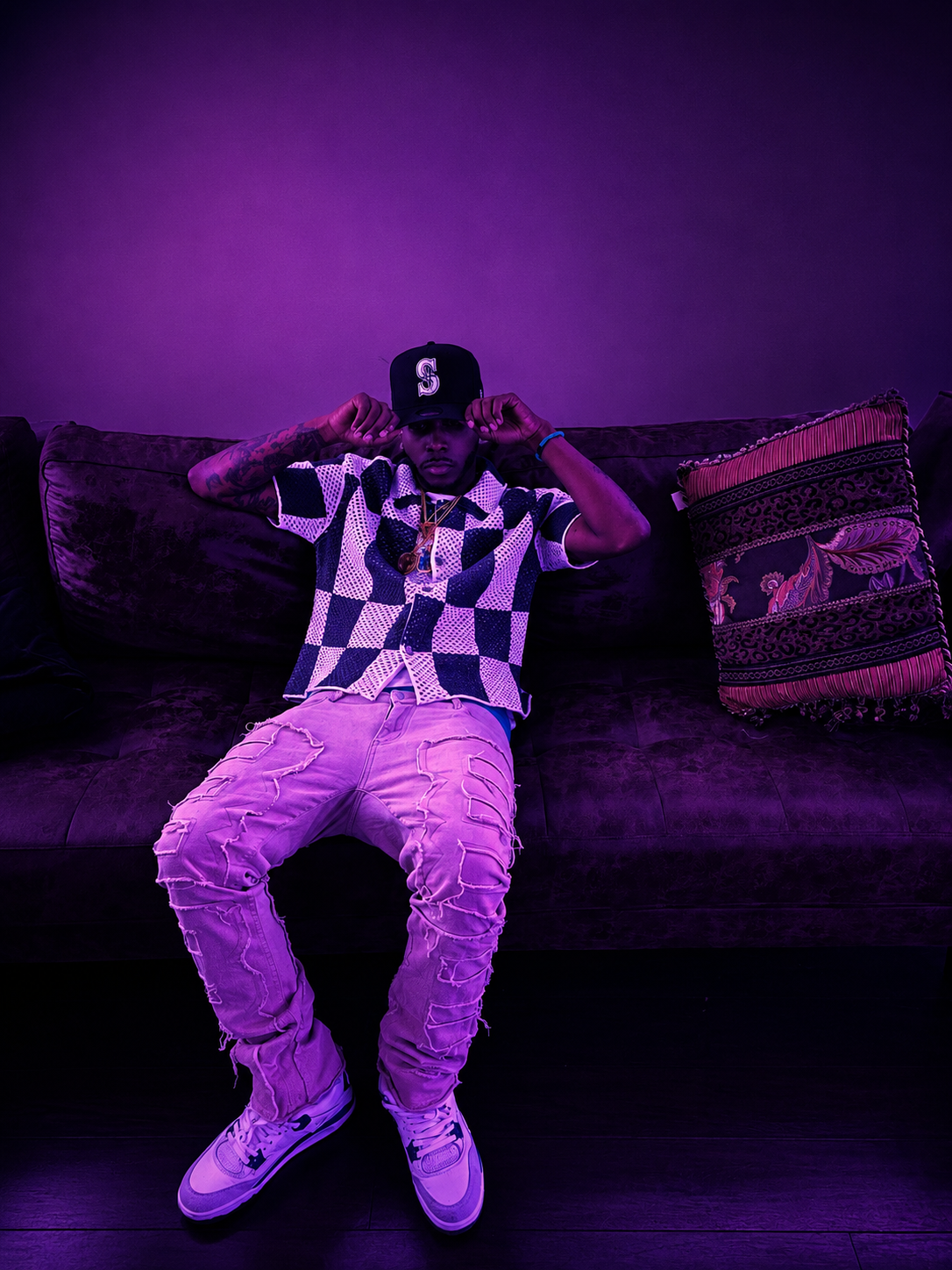A young man sitting on a dark couch in a room bathed in purple lighting. He is wearing a black baseball cap, a patterned short-sleeve shirt, light purple ripped jeans, white sneakers, and jewelry. One hand is on his cap, and he is looking at the camera.