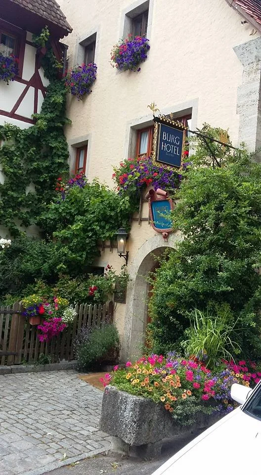 A charming European street scene featuring a hotel entrance with flower boxes on windows, lush greenery, and vibrant colorful flowers in a stone planter. The building has a white stucco facade with a mix of traditional and alpine style architecture.