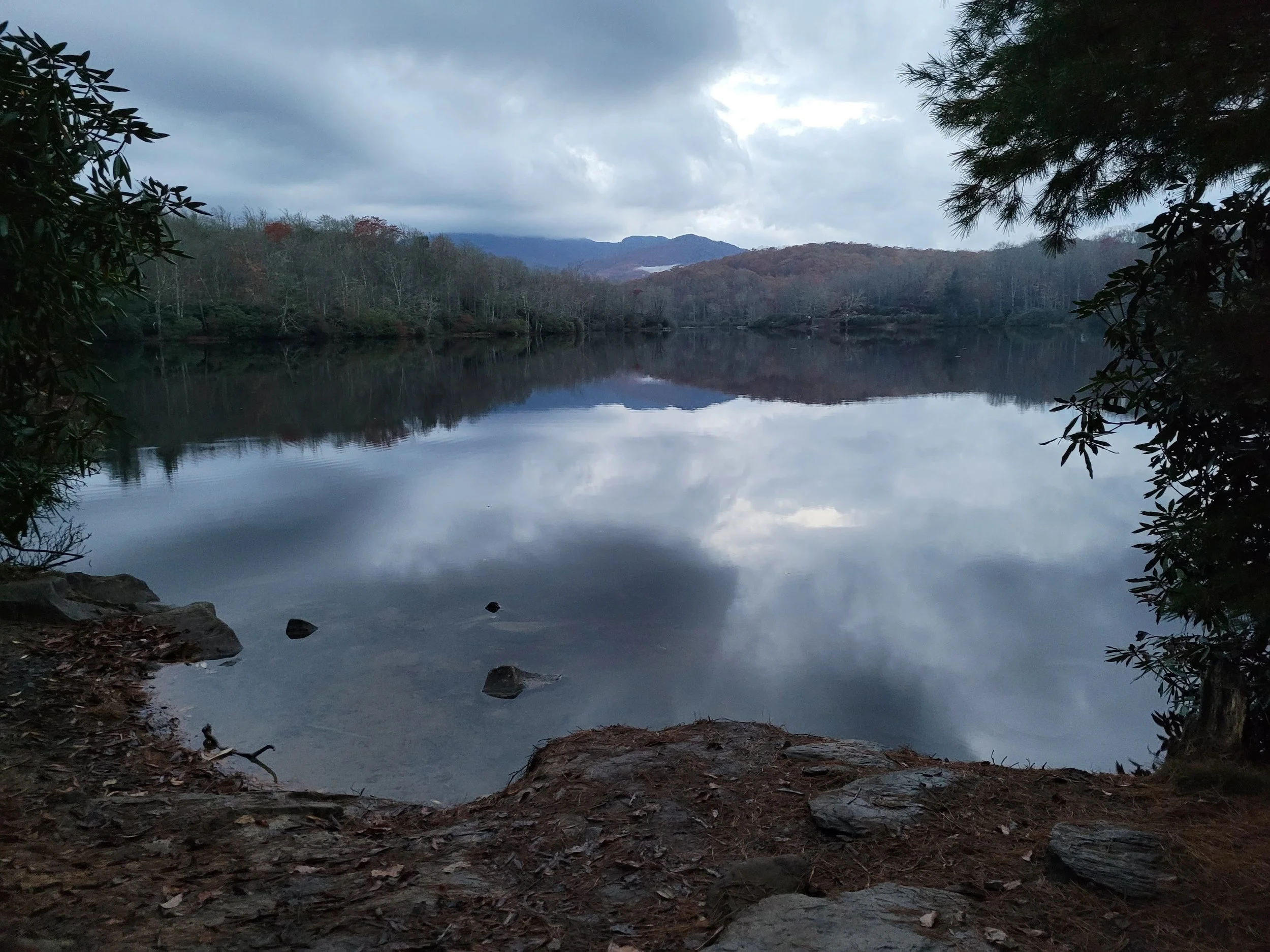 A calm lake surrounded by trees and distant hills under a cloudy sky.