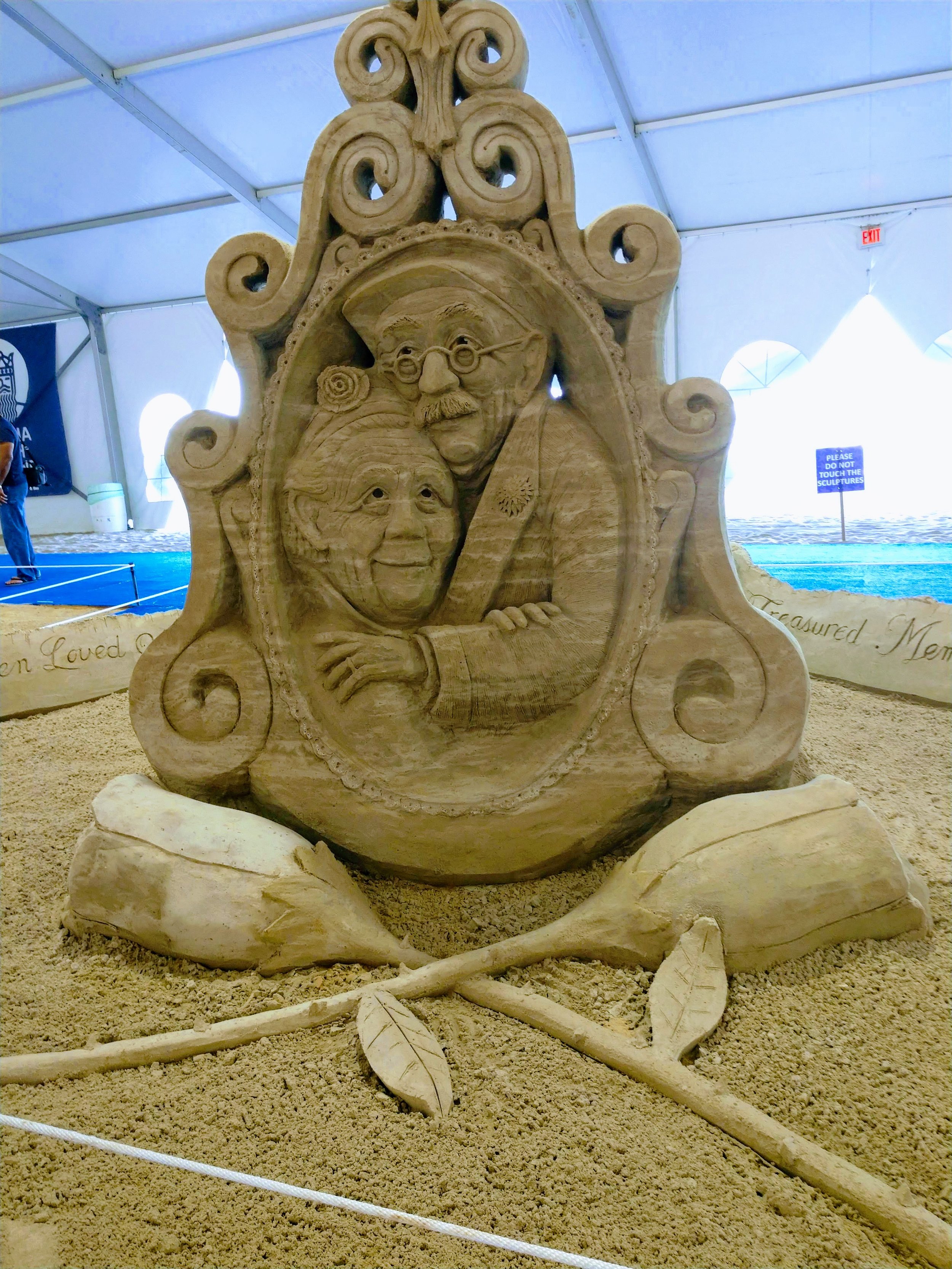 Sand sculpture featuring a framed portrait of a woman and a bearded man with glasses, surrounded by decorative scrollwork and sculptures of leaves and branches.