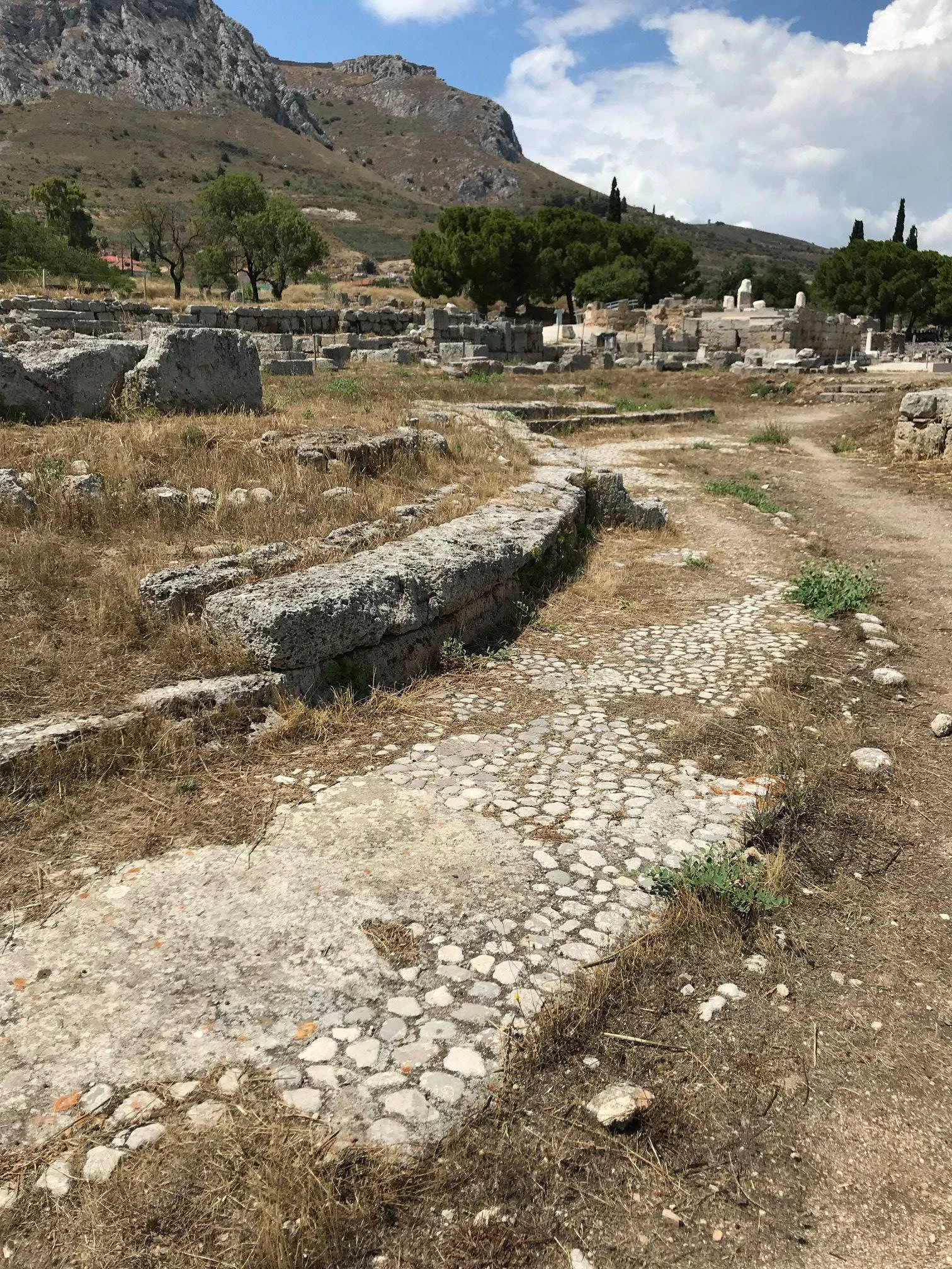 Ancient ruins with stone pathways and scattered large stones, set in a dry landscape with sparse grass and trees, mountains in the background under a partly cloudy sky.