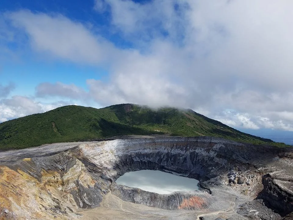 A volcanic crater with a lake inside, surrounded by volcanic rocks and a green mountain in the background, under a partly cloudy sky.