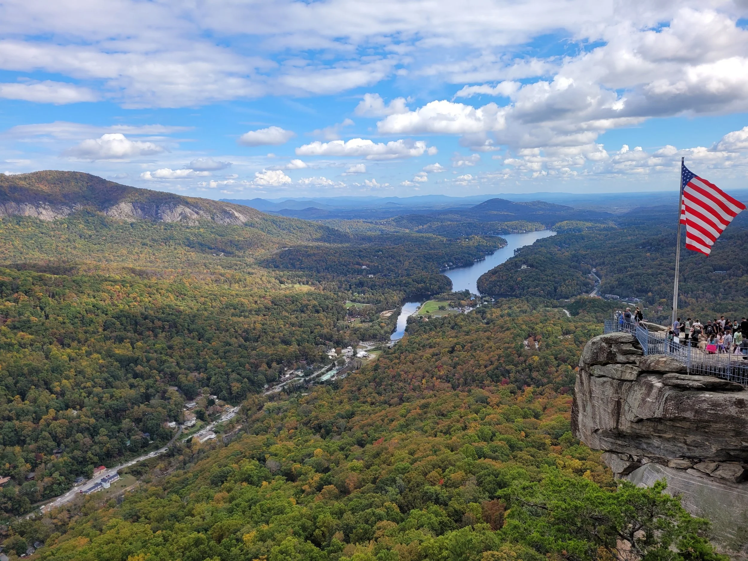 A scenic mountain landscape seen from a high vantage point with a partly cloudy sky, a winding river, and a group of people standing on a viewing platform near a flagpole with an American flag.