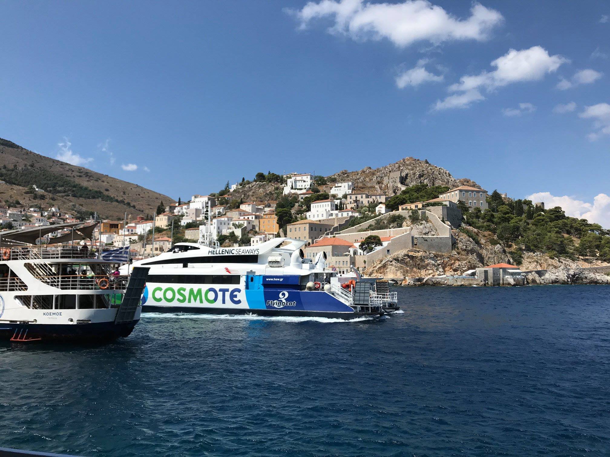 View of a harbor with boats, a hillside with houses, and a blue sky with clouds.