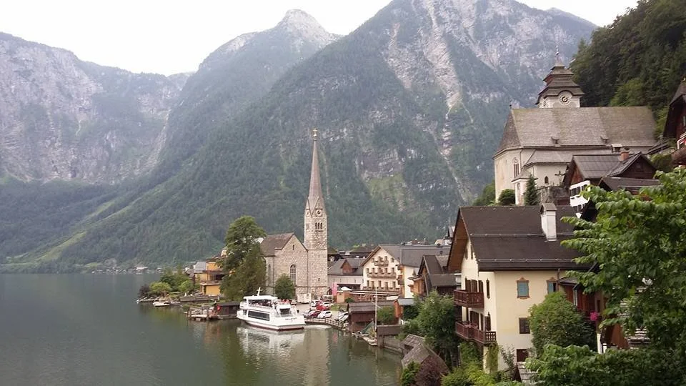 A picturesque lakeside village surrounded by steep, forested mountain cliffs with a church featuring a tall steeple, nestled among traditional buildings and boats docked at the water's edge.