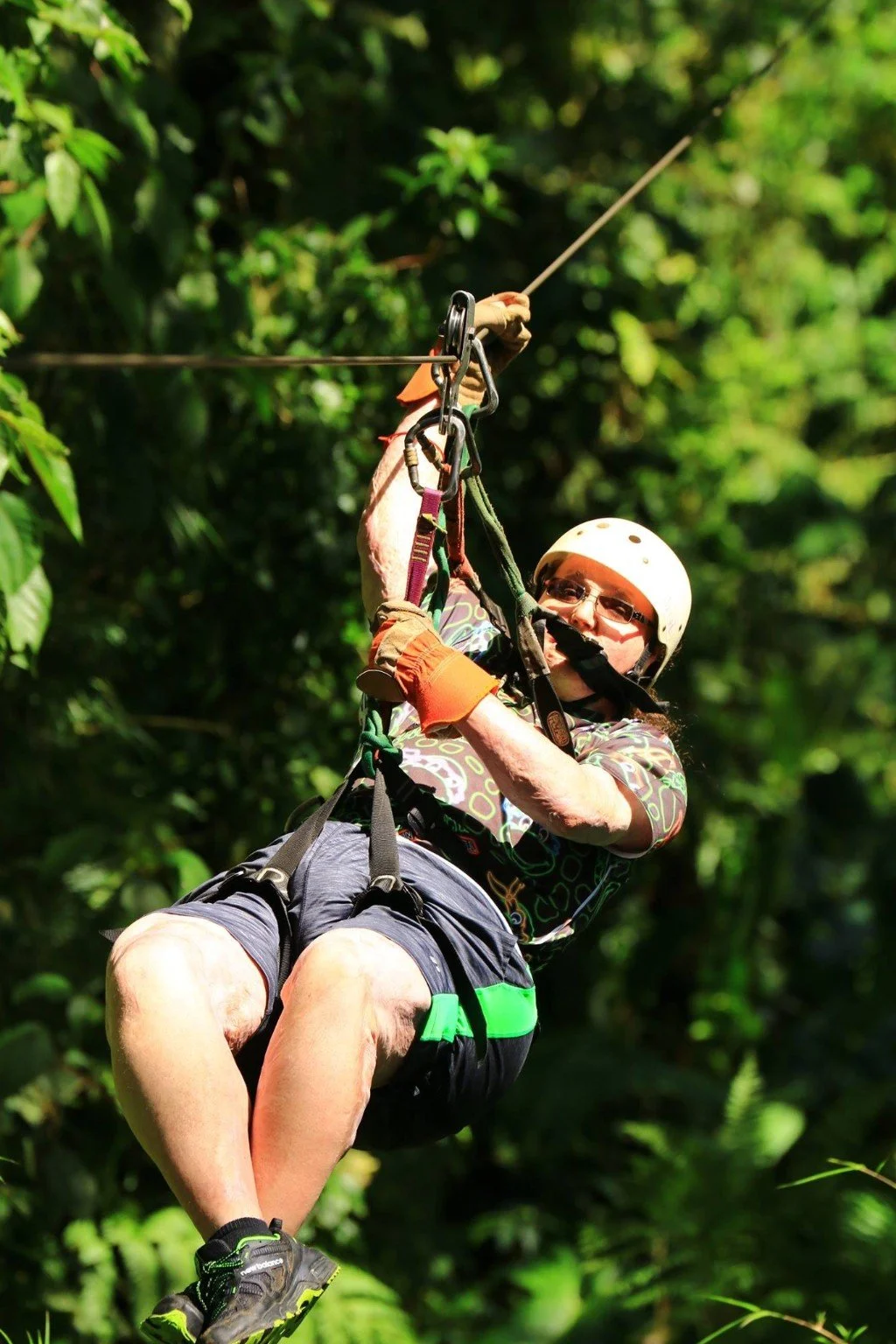 A man wearing safety gear, including a helmet and gloves, is zip-lining through a forest with lush green foliage.