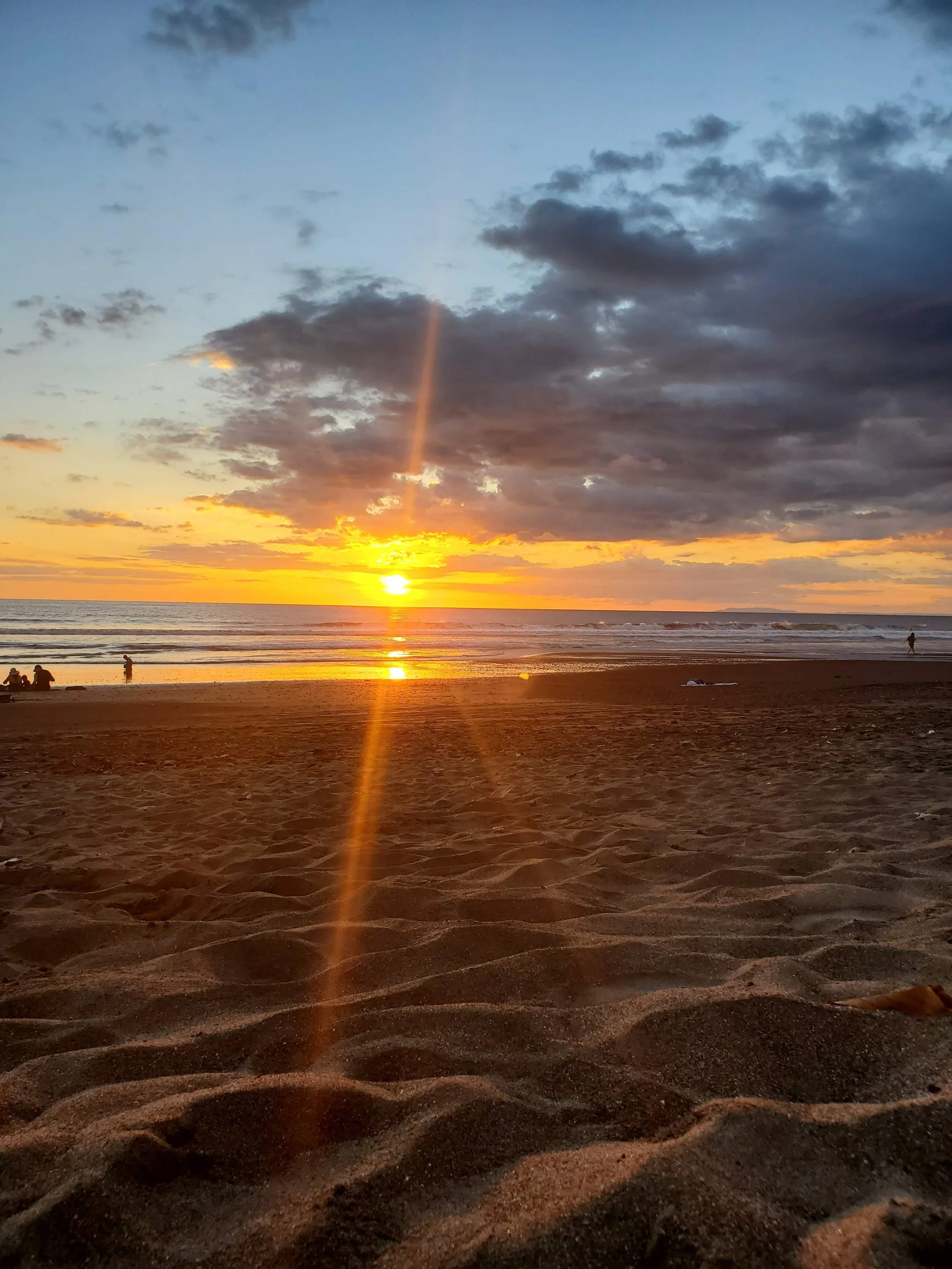 Sunset over the ocean with a rainbow-like light beam in the sky, sandy beach in the foreground, some trees at the bottom corners.