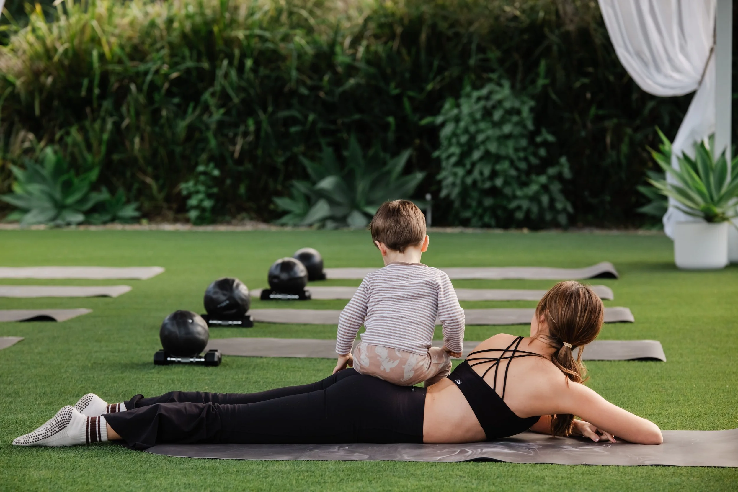 Outdoor mat mums and bubs pilates classA woman in black workout clothes lying on her stomach on a yoga mat on a grassy outdoor area, with child sitting on her back, surrounded by pilates mats, equipment and a lush green hedge in the background.