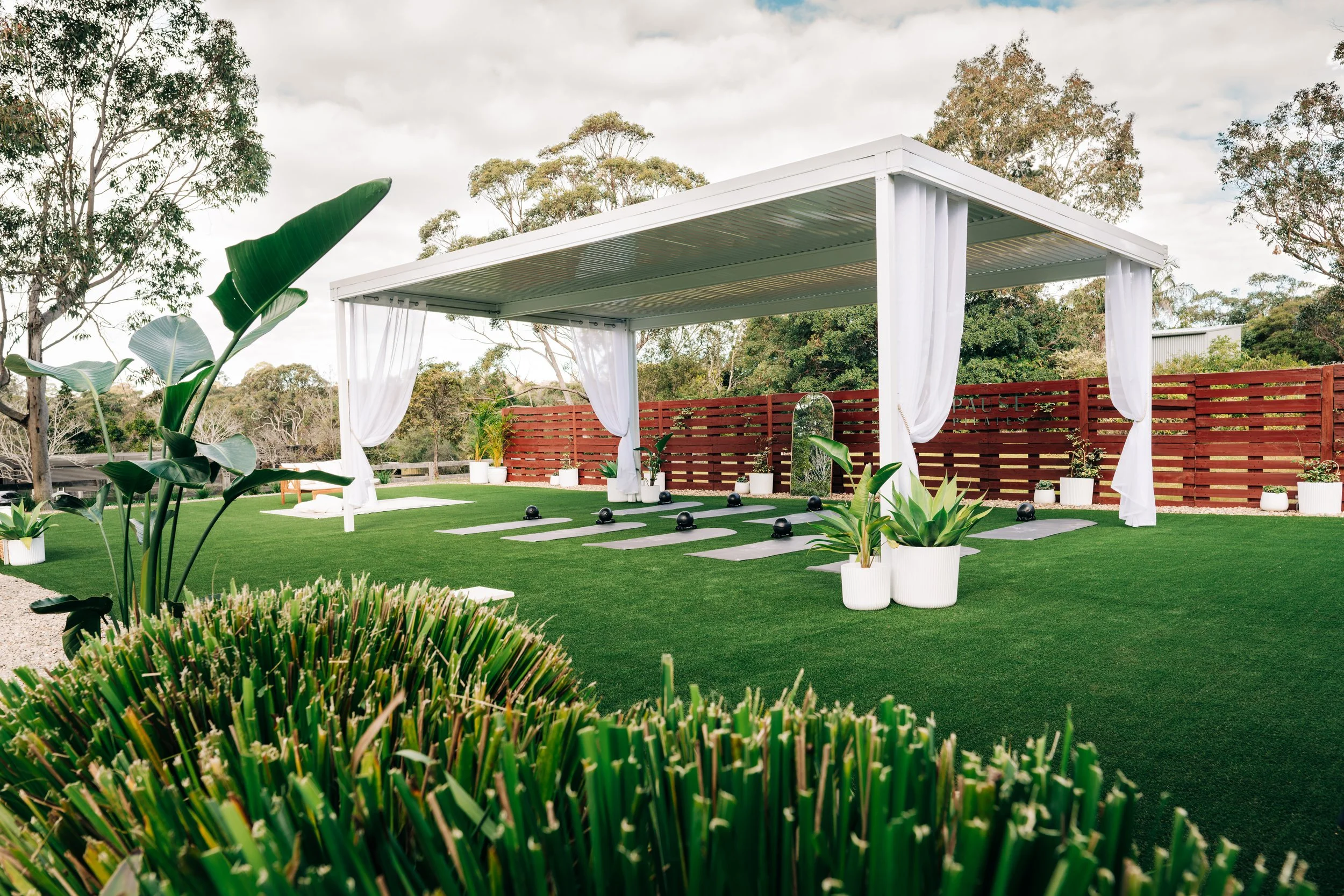 Outdoor yoga space with a white canopy, white curtains, yoga mats, black blocks, surrounded by potted plants and greenery, with trees in the background.