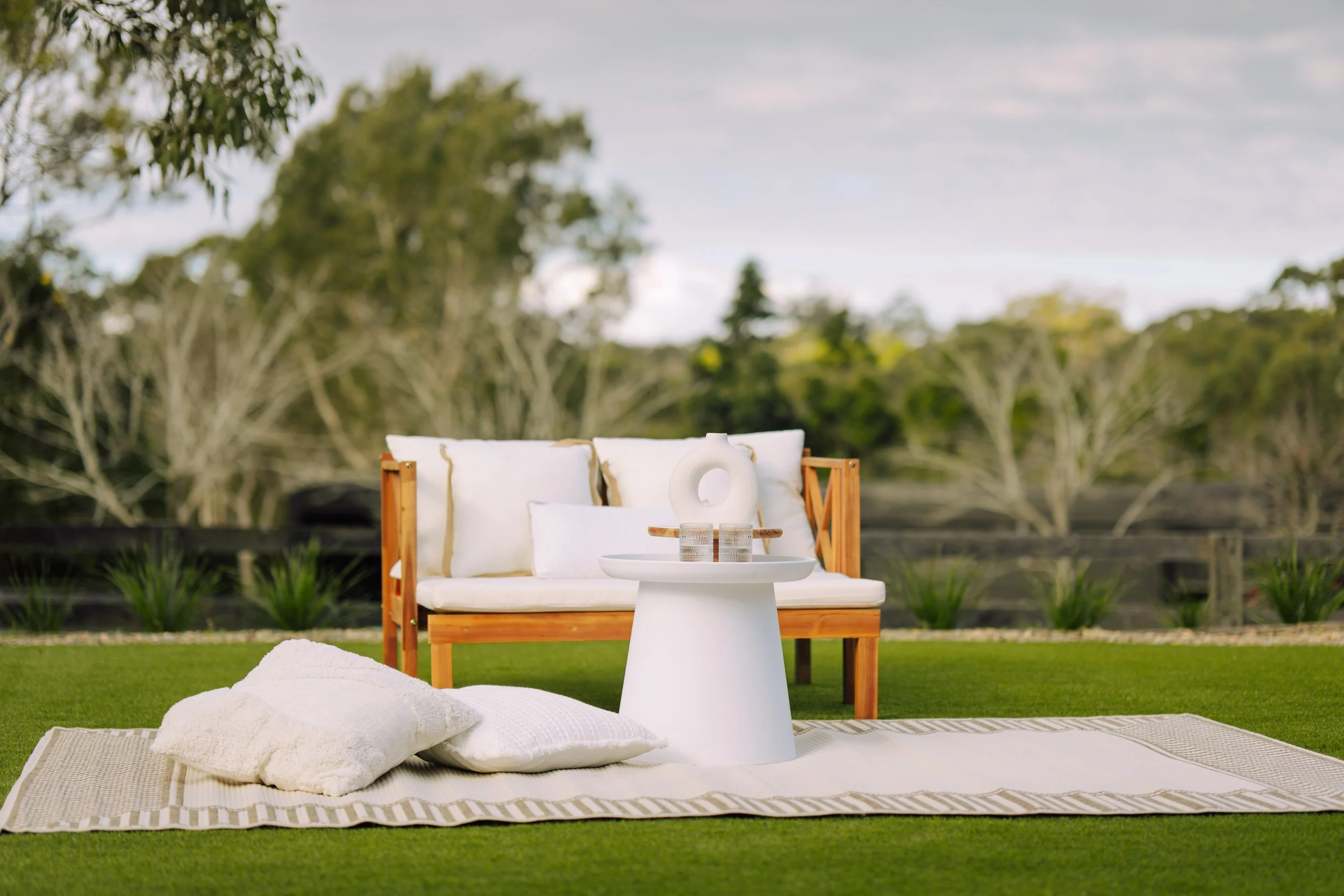 Outdoor mat pilates studio in Sydney featuring outdoor lounge area with a wooden sofa and white cushions, a white side table with glass cups and a decorative item, set on a beige rug on green grass, with trees and a cloudy sky in the background. 