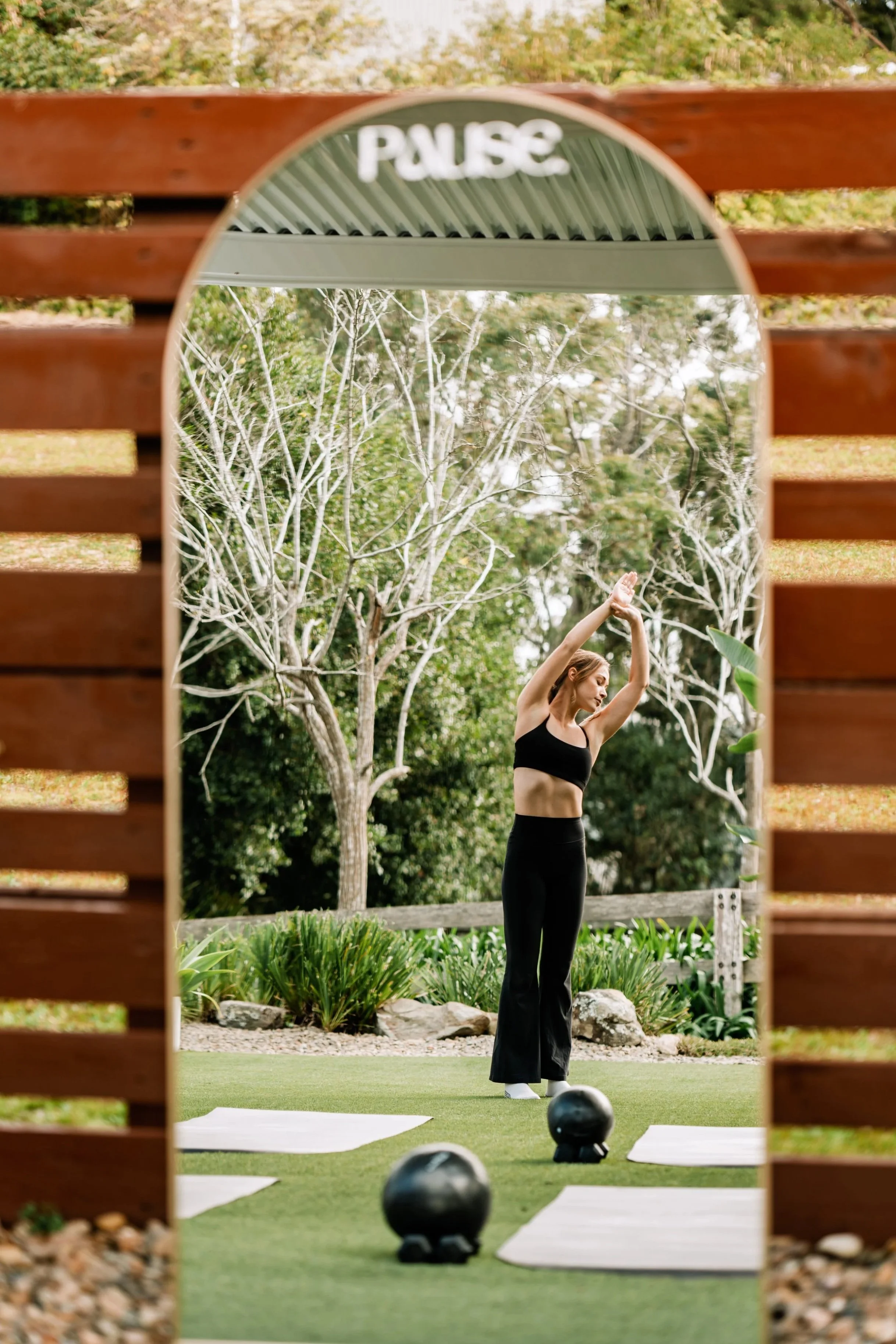 Woman practicing pilates outdoors in an outdoor boutique pilates studio, seen through an oval-shaped mirror. Aesthetic pilates photo.