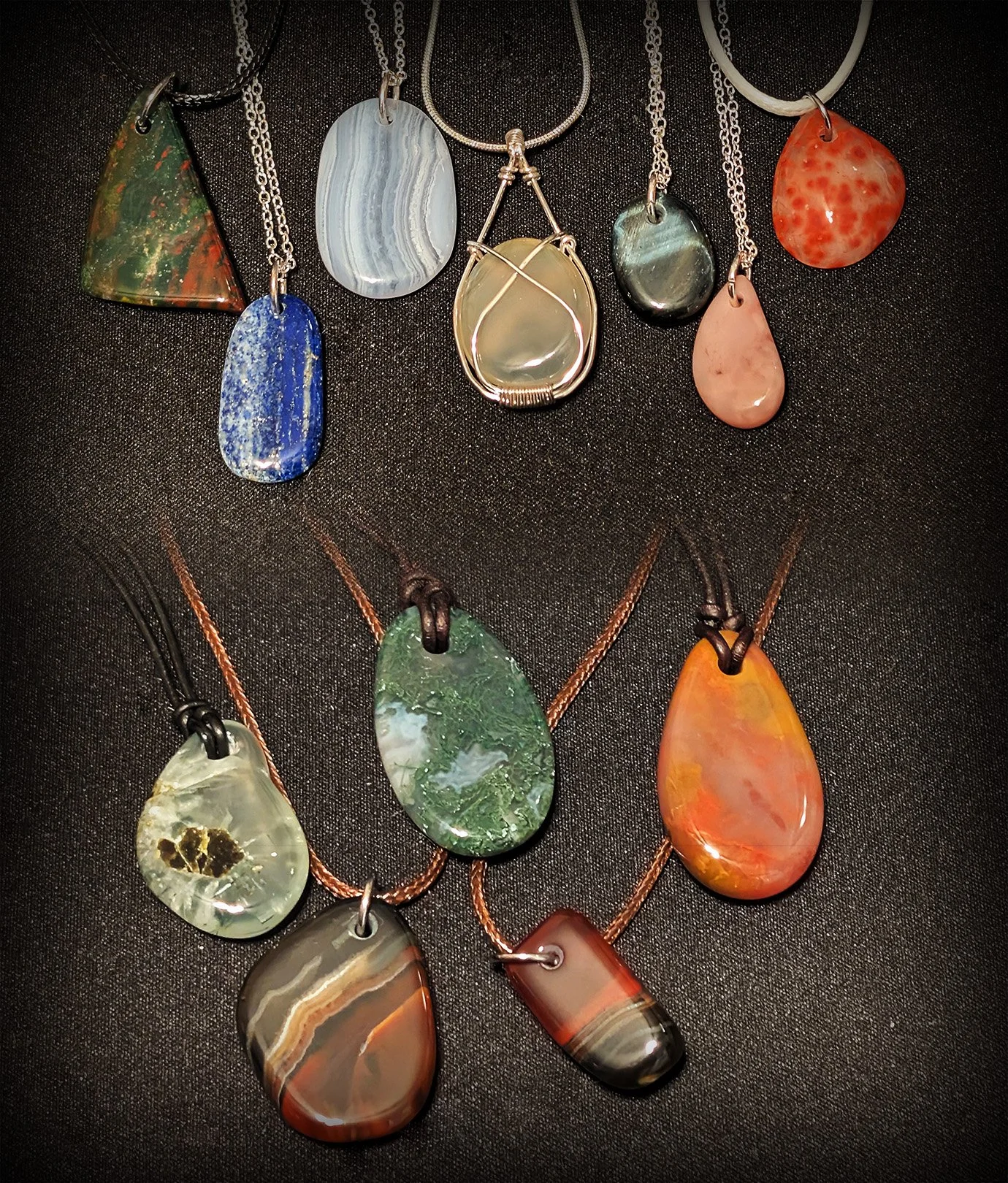 Various colorful, polished stone pendants with silver and leather cords laid on black fabric.