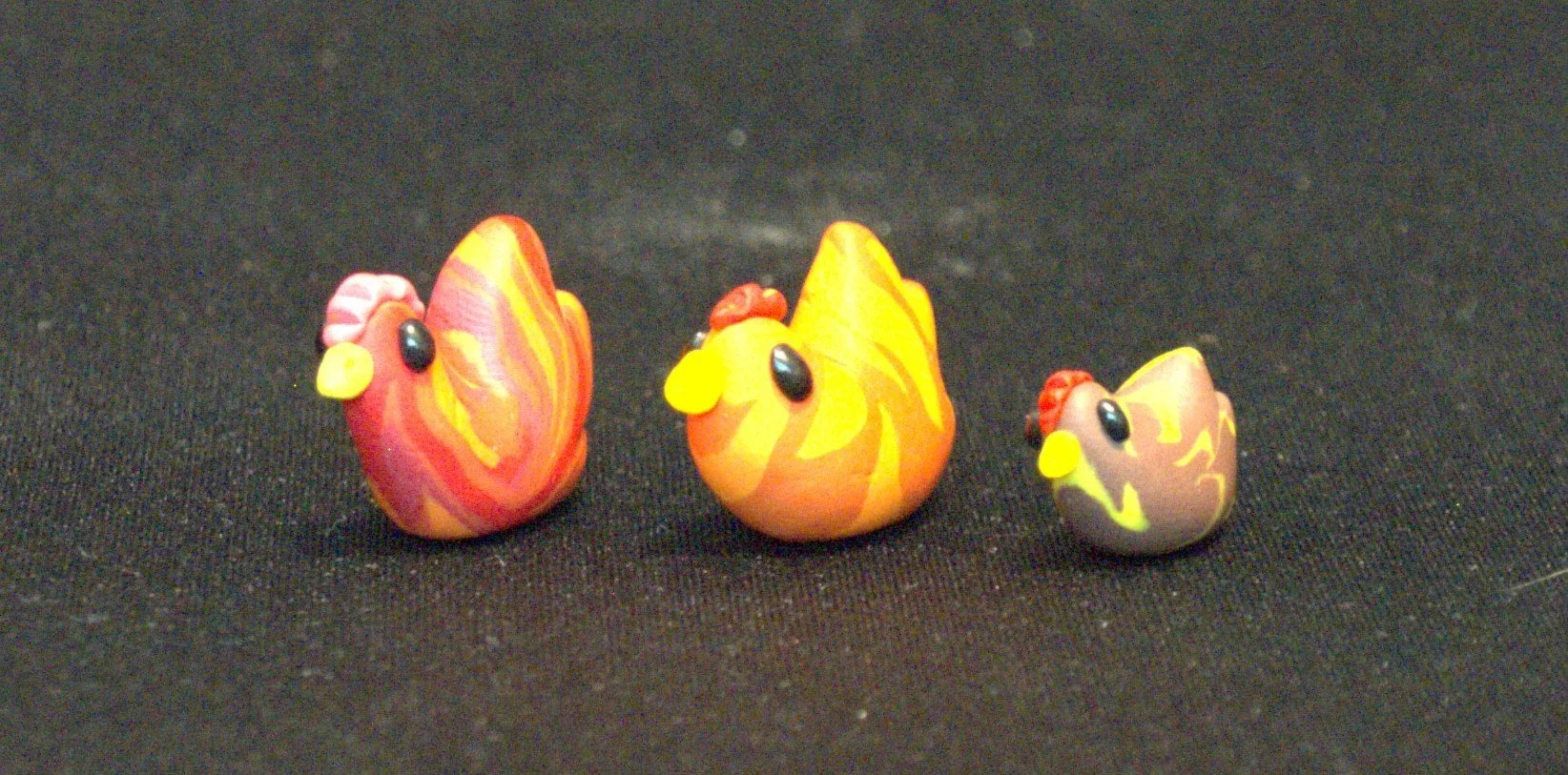 Three colorful small chicken figurines made of clay, arranged in a row on a black surface.