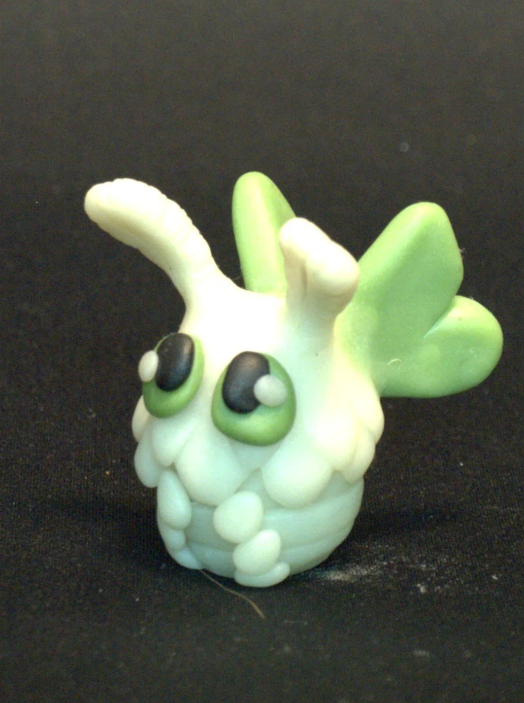 A small, white, cartoonish figurine resembling a chubby moth with big green eyes and wings, against a black background.