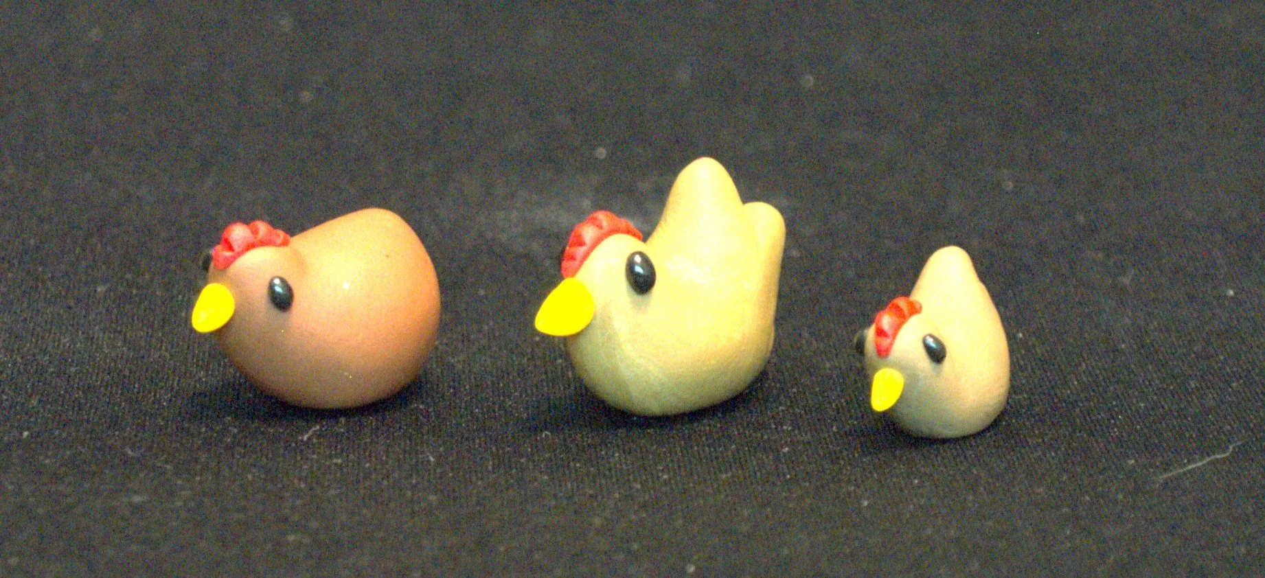 Three small chicken figurines with yellow beaks, red combs, black eyes, and bodies colored brown, golden brown, and tan, positioned on a black surface.