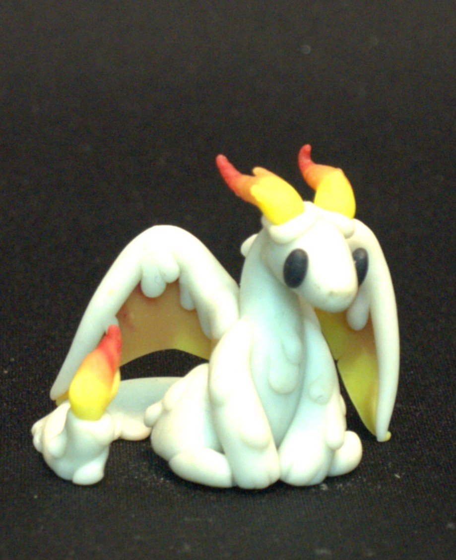 A small, cute figurine of a dragon modeled after a candle with a white body, yellow wings, orange-tipped horns, and a fire on the tip of the tail, sitting on a black surface.