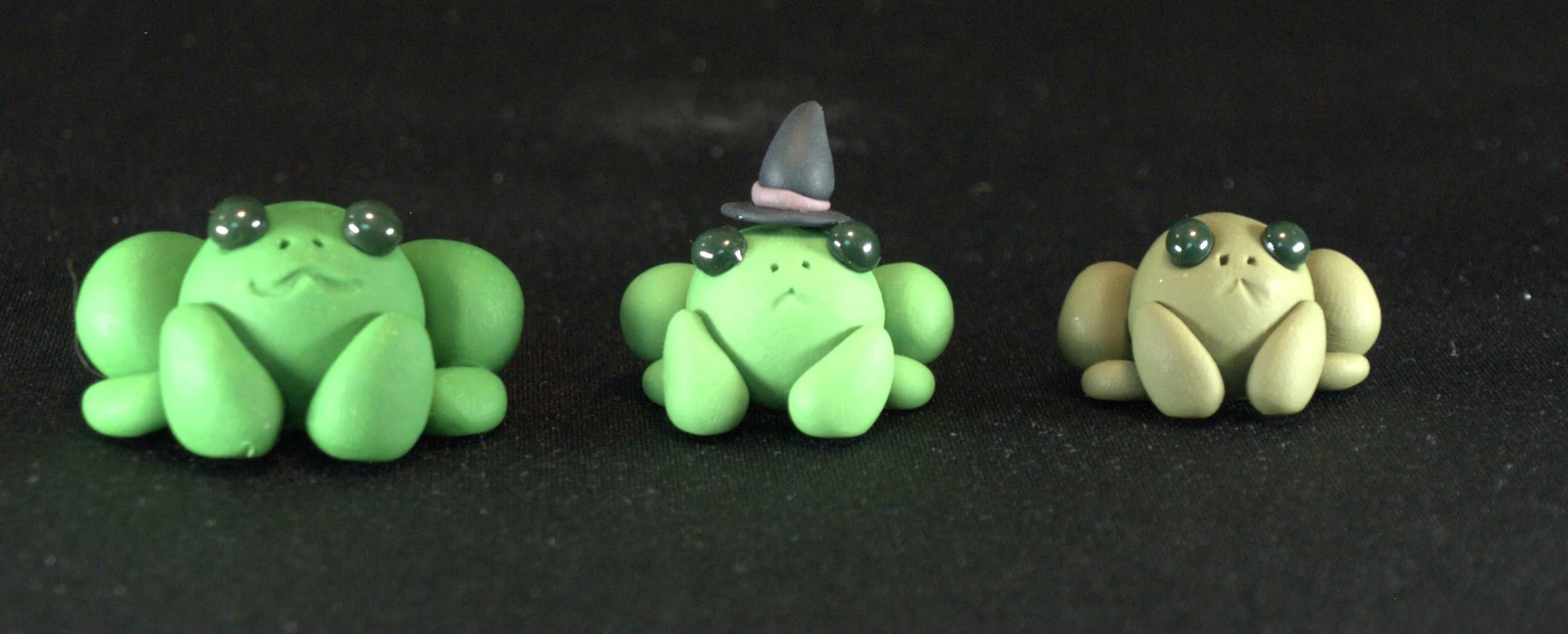 Three small frog figurines made of green and beige material with large black eyes, sitting on a black surface. The middle frog is wearing a pointed gray witch hat.