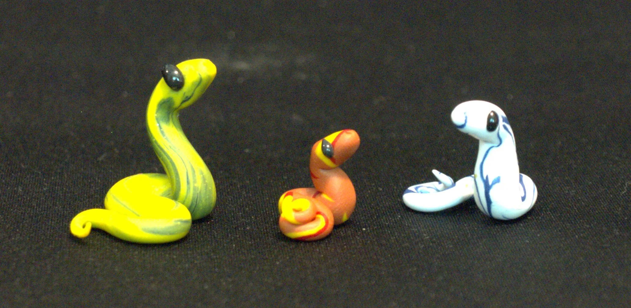 Three colorful clay snake figurines on a black background, each with distinct colors and patterns. The first is a swirl of yellow and blue, the second is a swirl of red and yellow, the third is marbled blue and white.