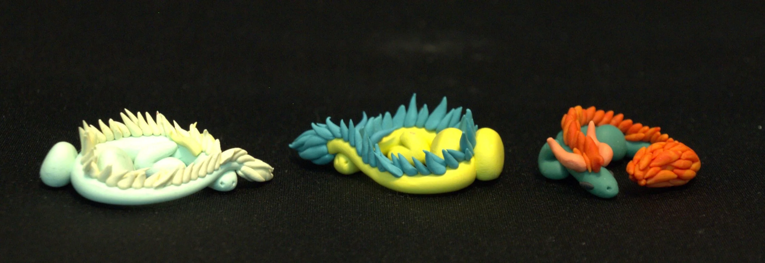 Three small eastern dragon figurines in light blue, yellow, and orange made of clay on a black surface.