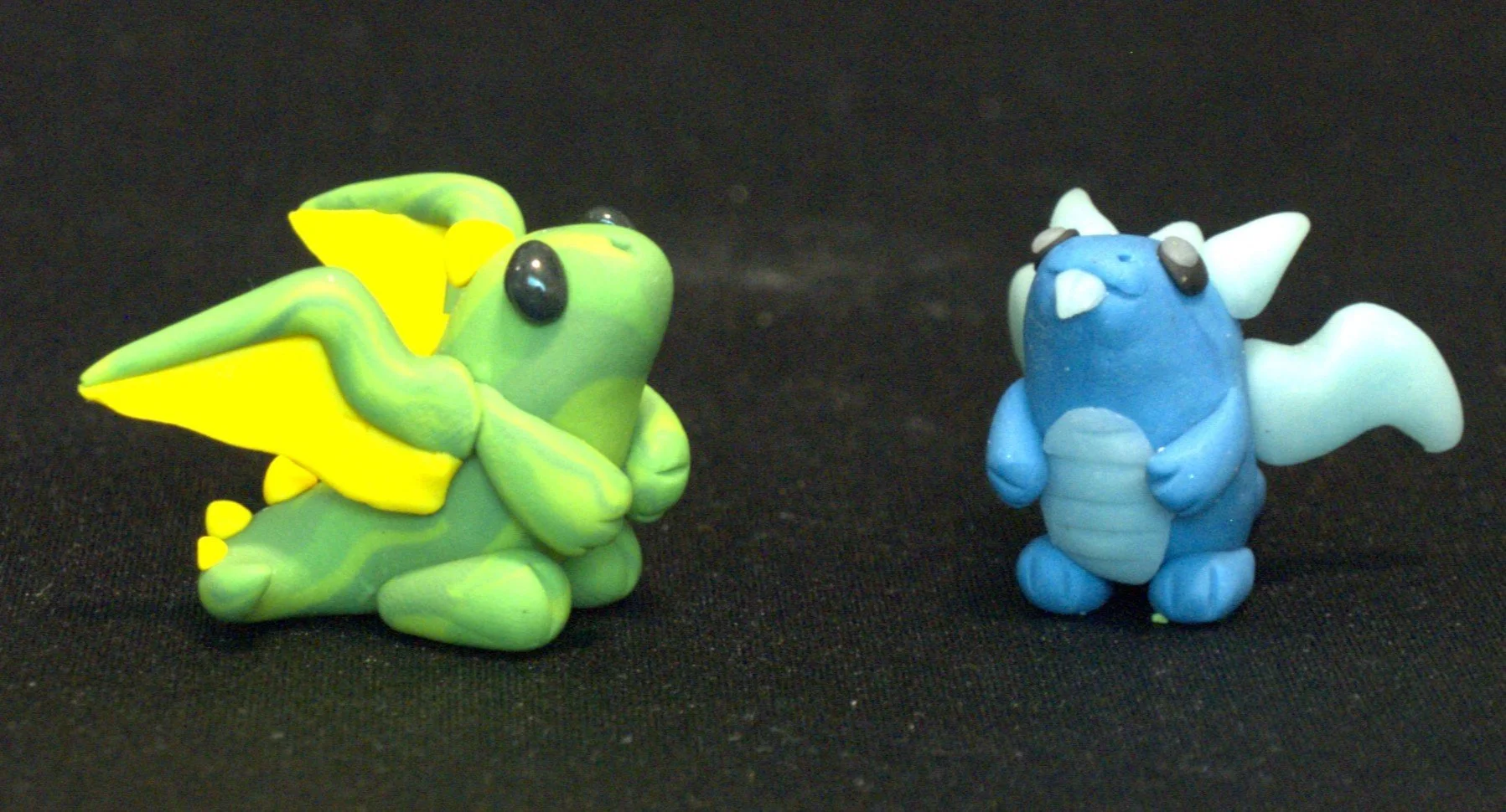 Two clay figurines of chibi styled dragons, the one on the left is green with yellow wings, and the one on the right is blue with a light blue belly and wings, placed on a black surface.