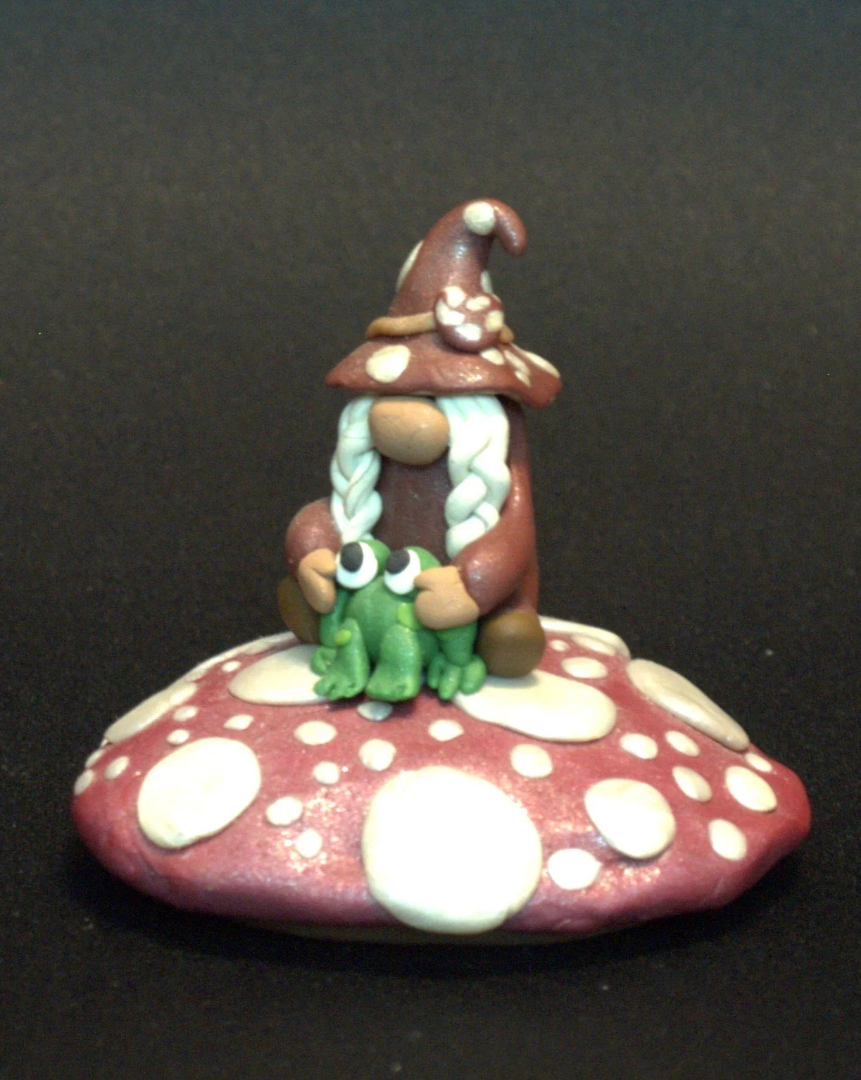 A figurine of a gnome with a red brown hat and white braids, sitting on a red and white spotted mushroom, holding a green frog with big eyes.