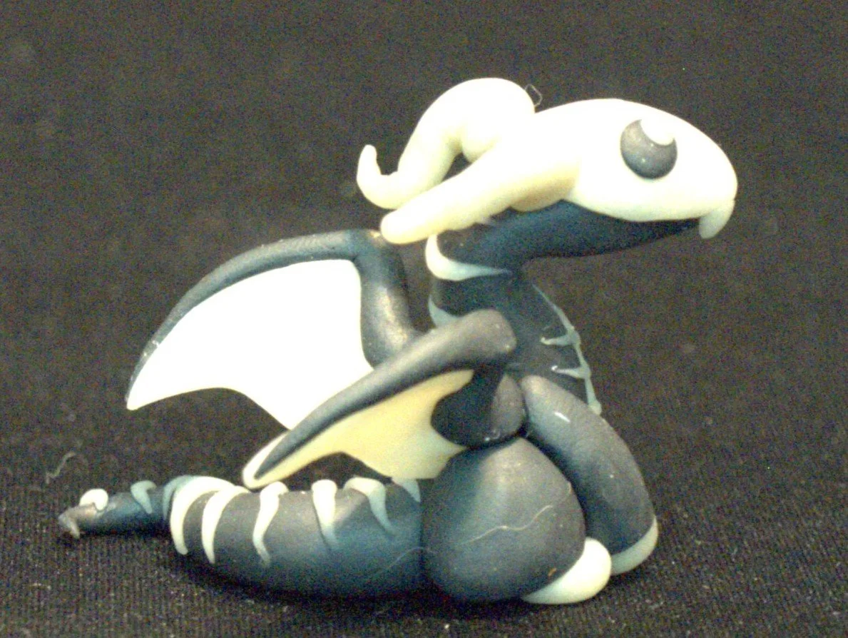 A handmade dragon figurine made of black and white modeling clay, featuring wings, a curled tail, and a skeleton pattern on its head and body with horns and large eyes.
