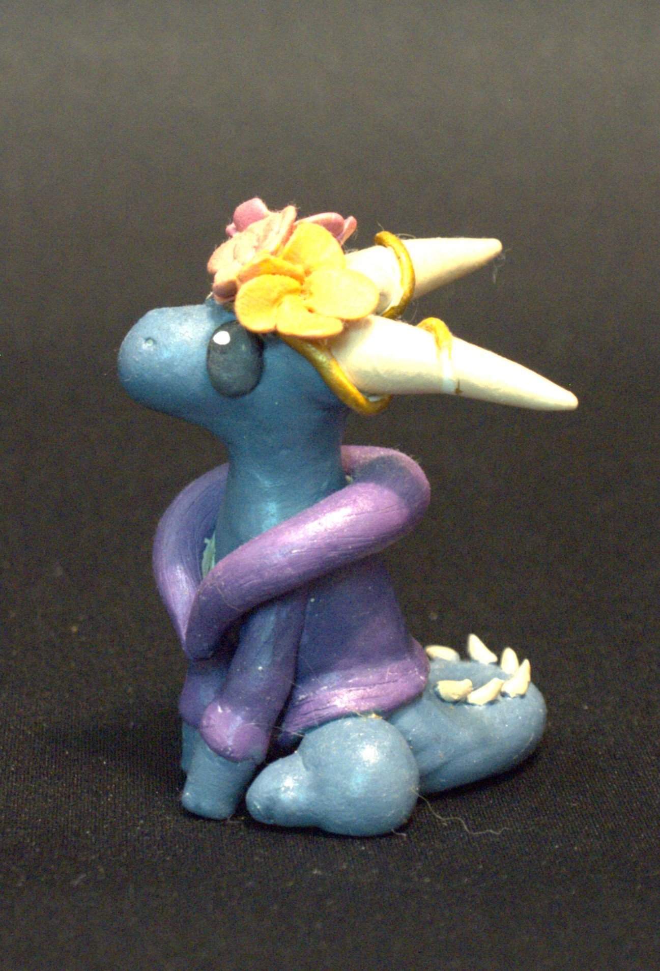 A decorative figurine of a dragon with a blue body, purple fluffy coat, and white horns, adorned with colorful flowers and yellow accents.