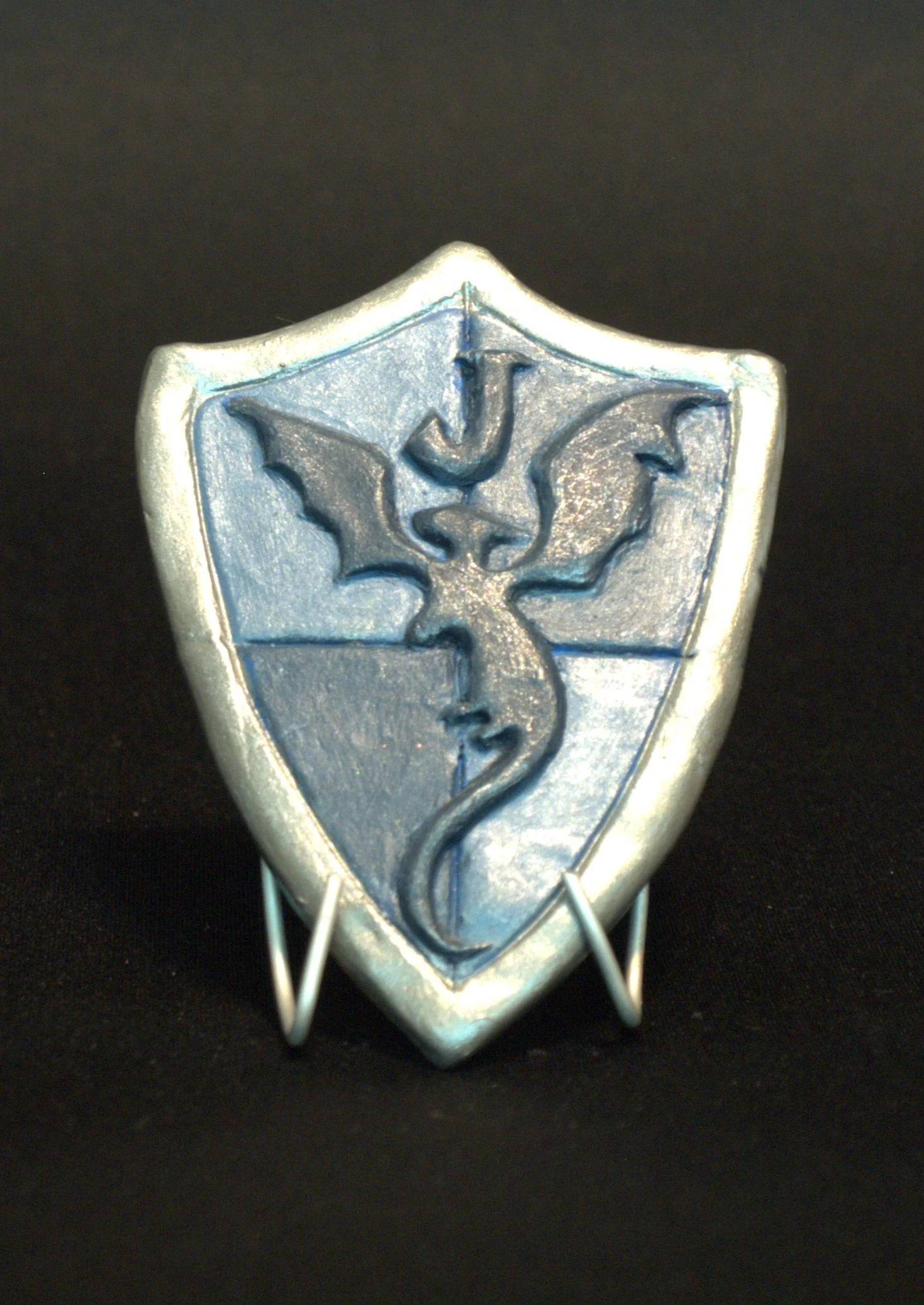 Close-up of a metallic shield-shaped badge with a dragon emblem, mounted on a black background with a wire stand at the bottom.