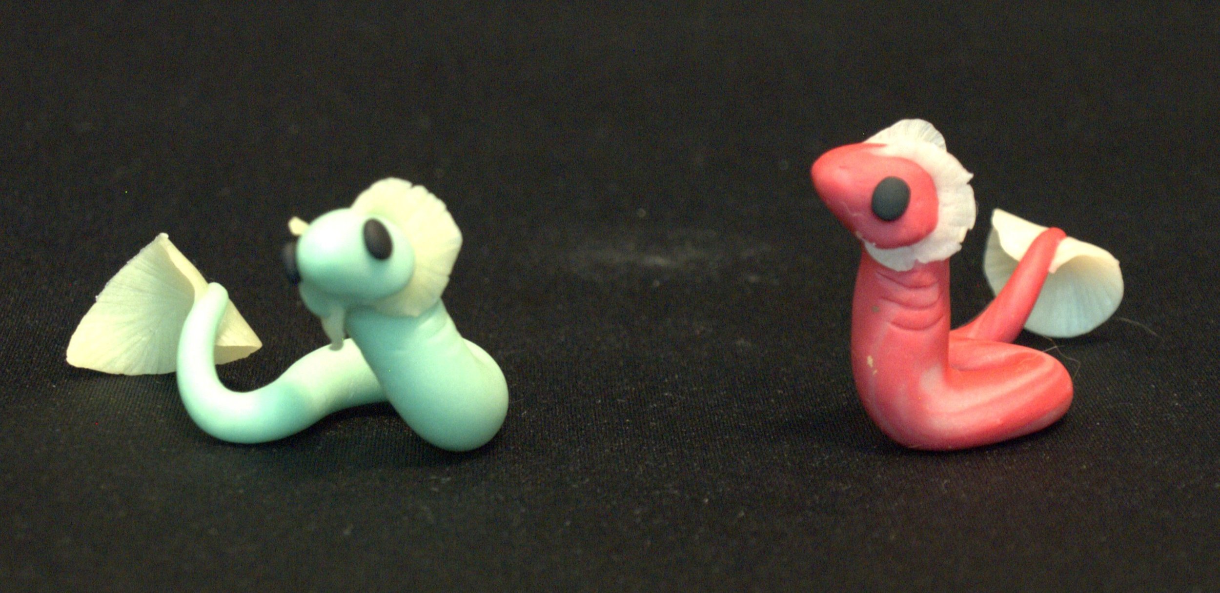 Two small, colorful, sea serpent figurines with ear frills and a fin on the end of the tail, one green and one pink, against a black background.