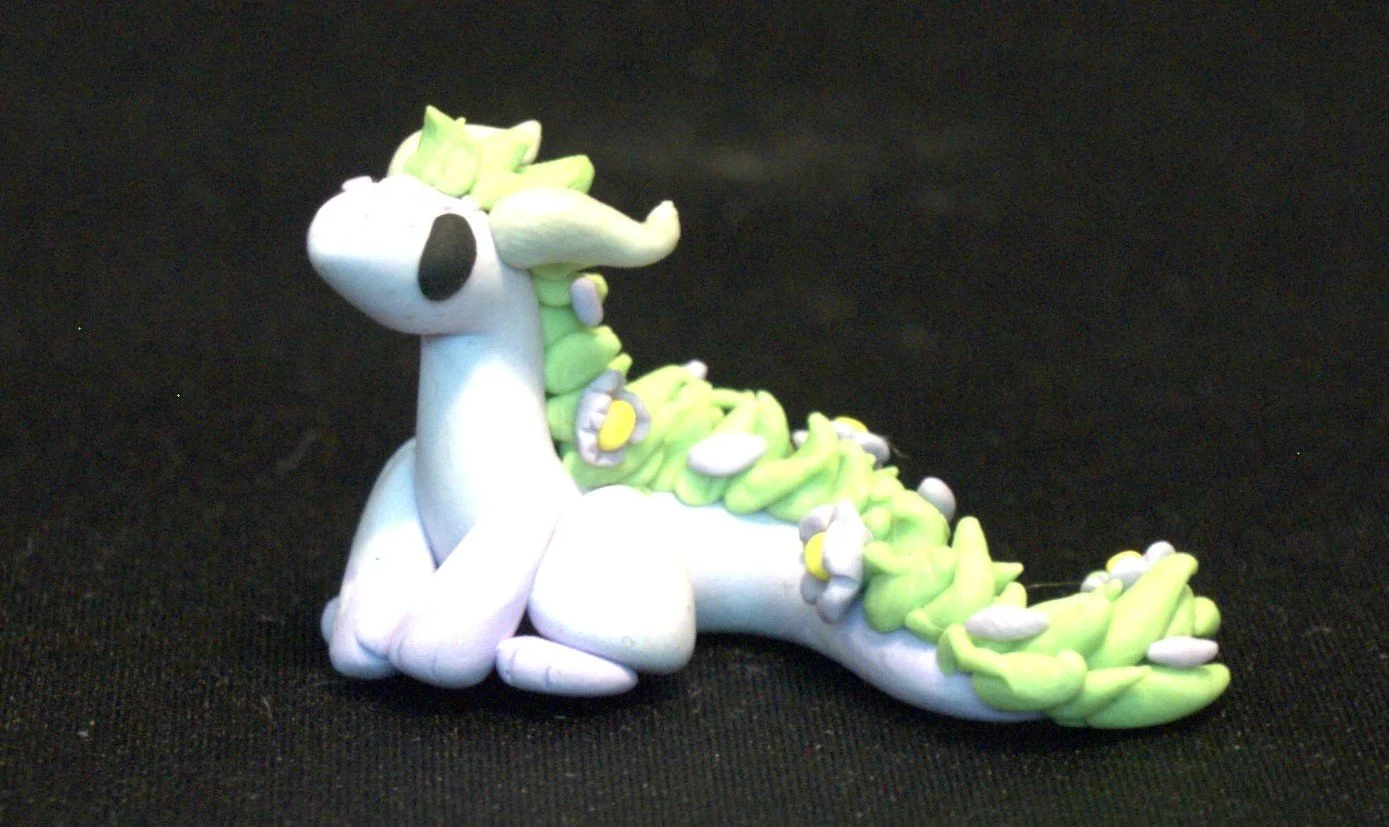 Clay model of a light lavender eastern dragon with lavender flowers in its light green mane, black eyes, sitting on a black surface.