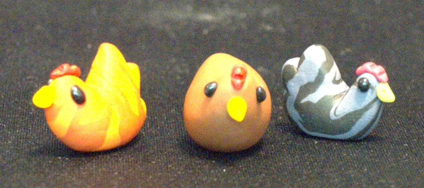 Three clay chickens, placed on a black surface. The first is a yellow chicken with a red comb, black eyes, and a yellow beak. The second is a brown chicken with a red comb, black eyes, and a yellow beak. The third chicken is gray and white with a red