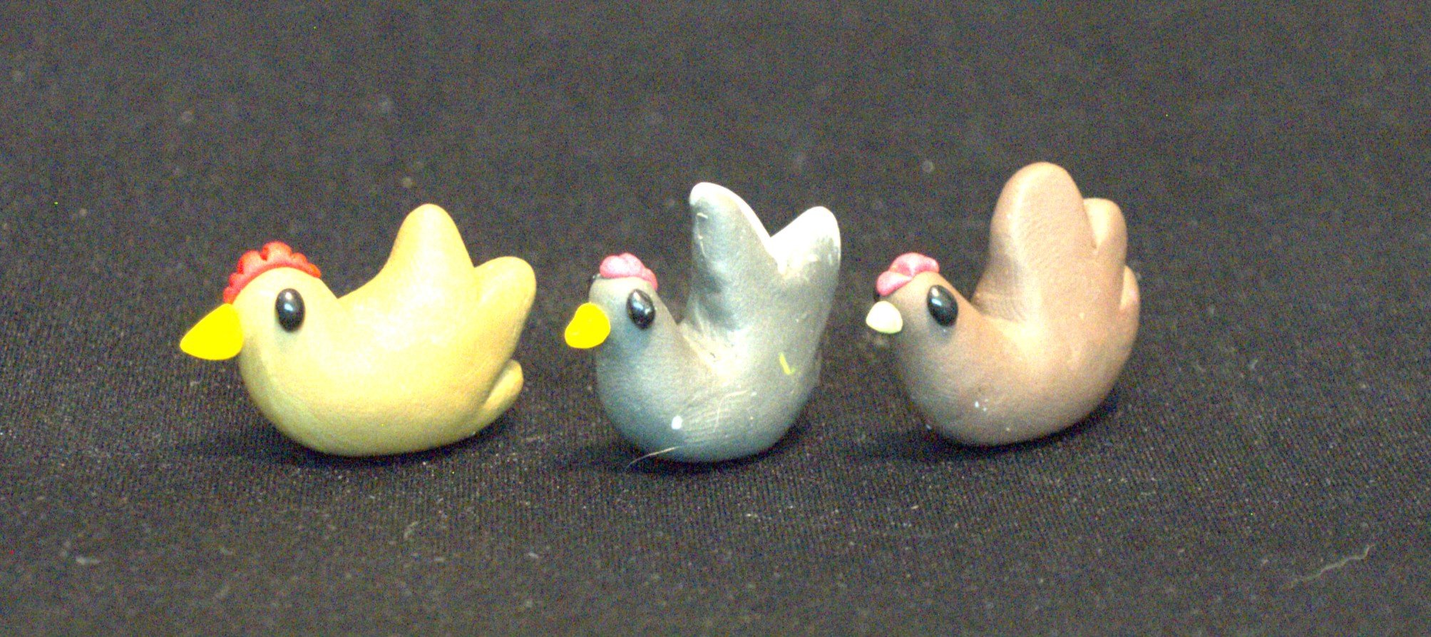 Three clay chicken figurines placed on a black surface, with yellow, gray, and light brown bodies, black eyes, yellow beaks, and red or pink combs.