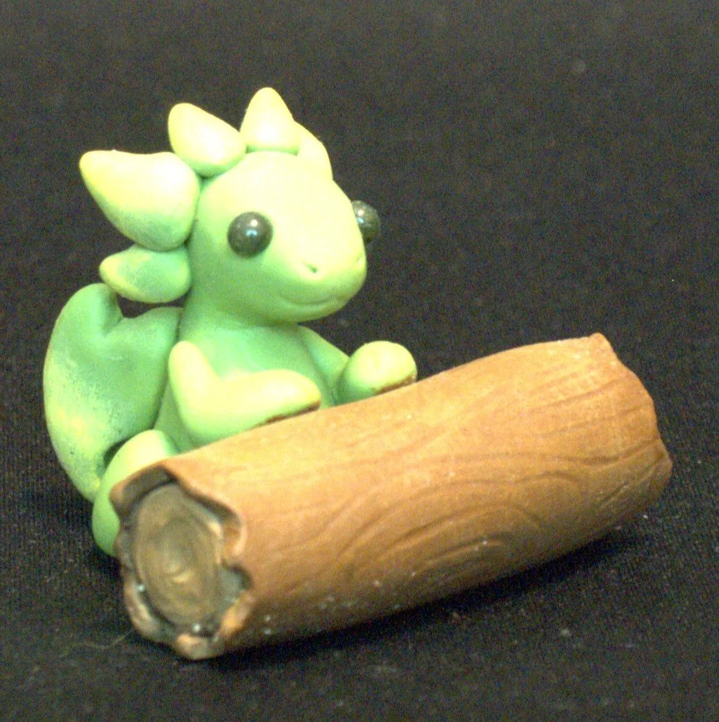 A small green dragon figurine with big black eyes, sitting with its front feet resting on a log, on a black background.