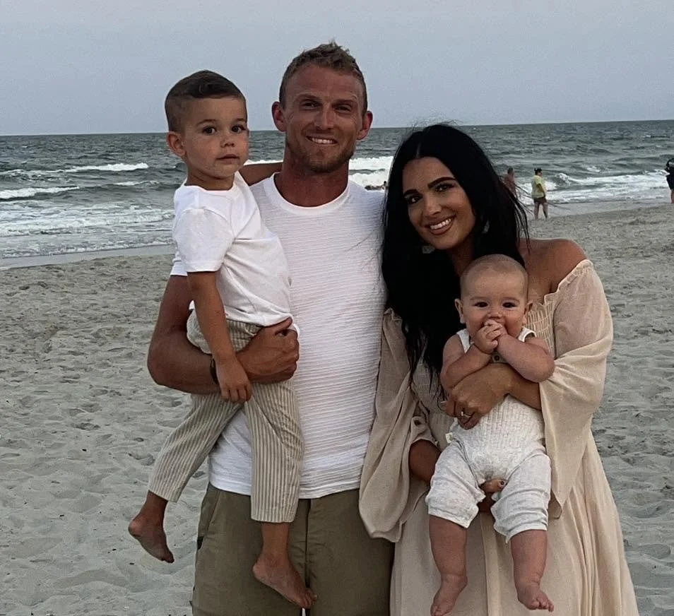 A smiling family of four on a beach with the ocean in the background, including a man holding a young boy, and a woman holding a baby.
