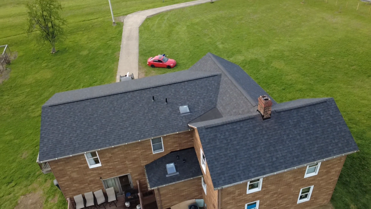 Aerial view of a two-story wooden house with a dark shingled roof, brick chimney, and small porch area, surrounded by a grassy yard with a paved walkway and a red car parked nearby.