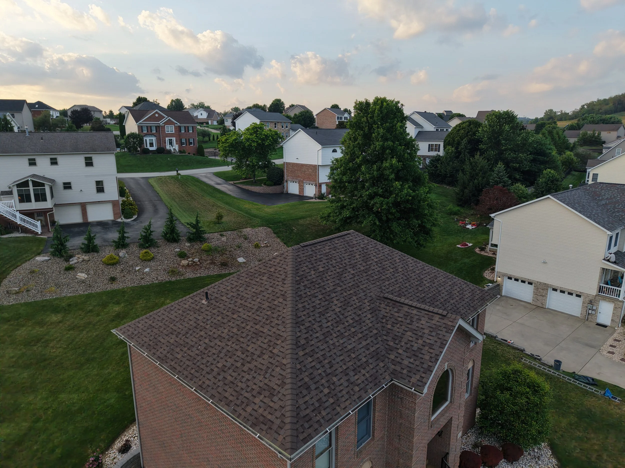 A suburban neighborhood with multiple houses, green lawns, large trees, and a sky with scattered clouds.