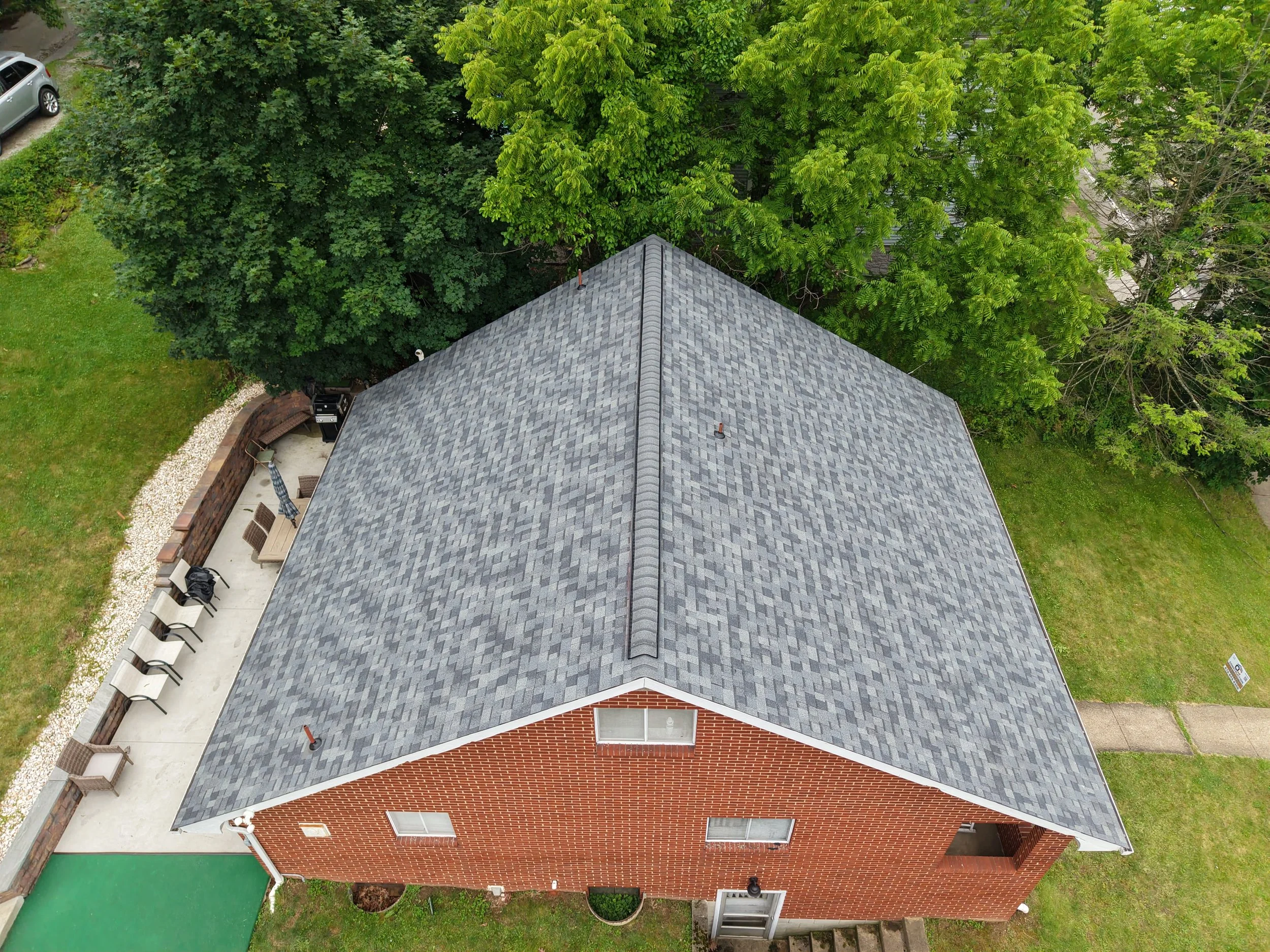 Aerial view of a house with a gray shingled roof surrounded by green trees, grass, and a small patio area with chairs and a grill.