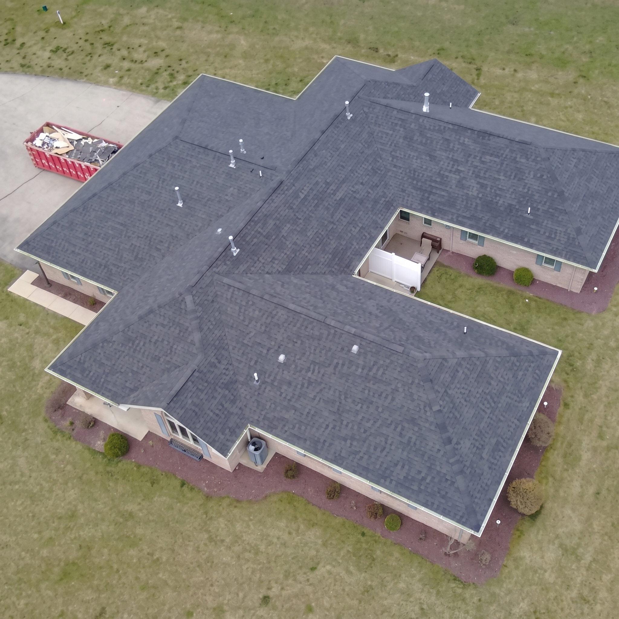 Aerial view of a house with a dark-shingled roof, multiple chimneys, and a backyard with brown grass. There is a red dumpster filled with construction debris on a paved driveway next to the house.