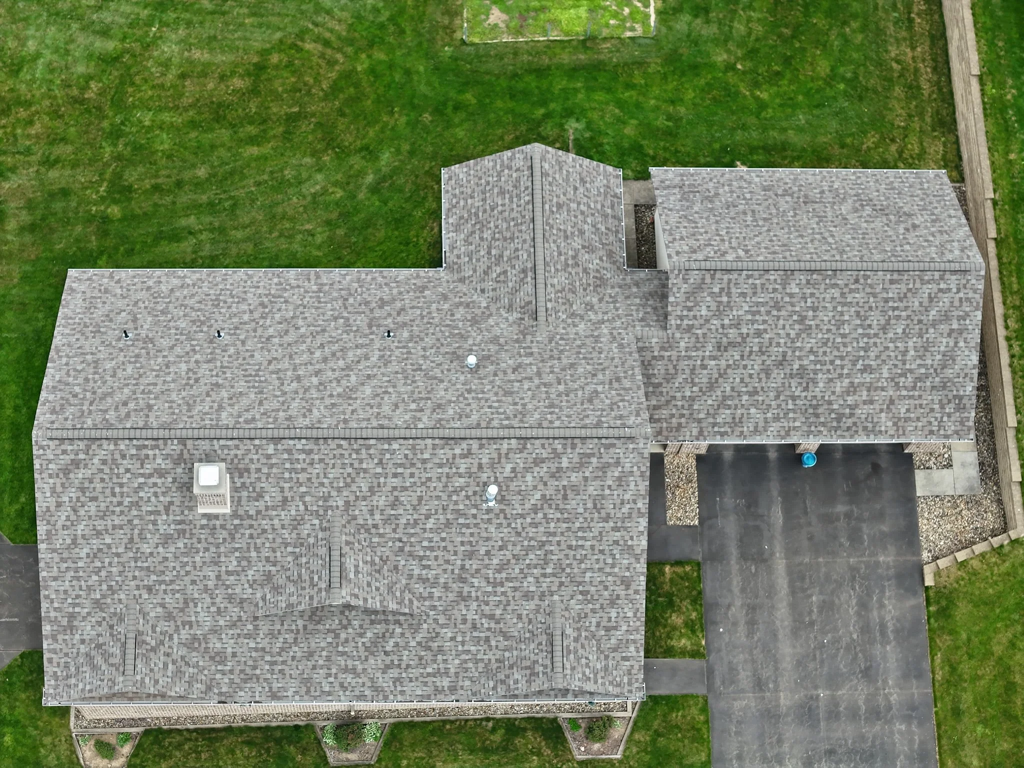 Aerial view of a house with a gray shingled roof, surrounded by a backyard with green grass, a paved driveway, and a wooden fence on the right side.