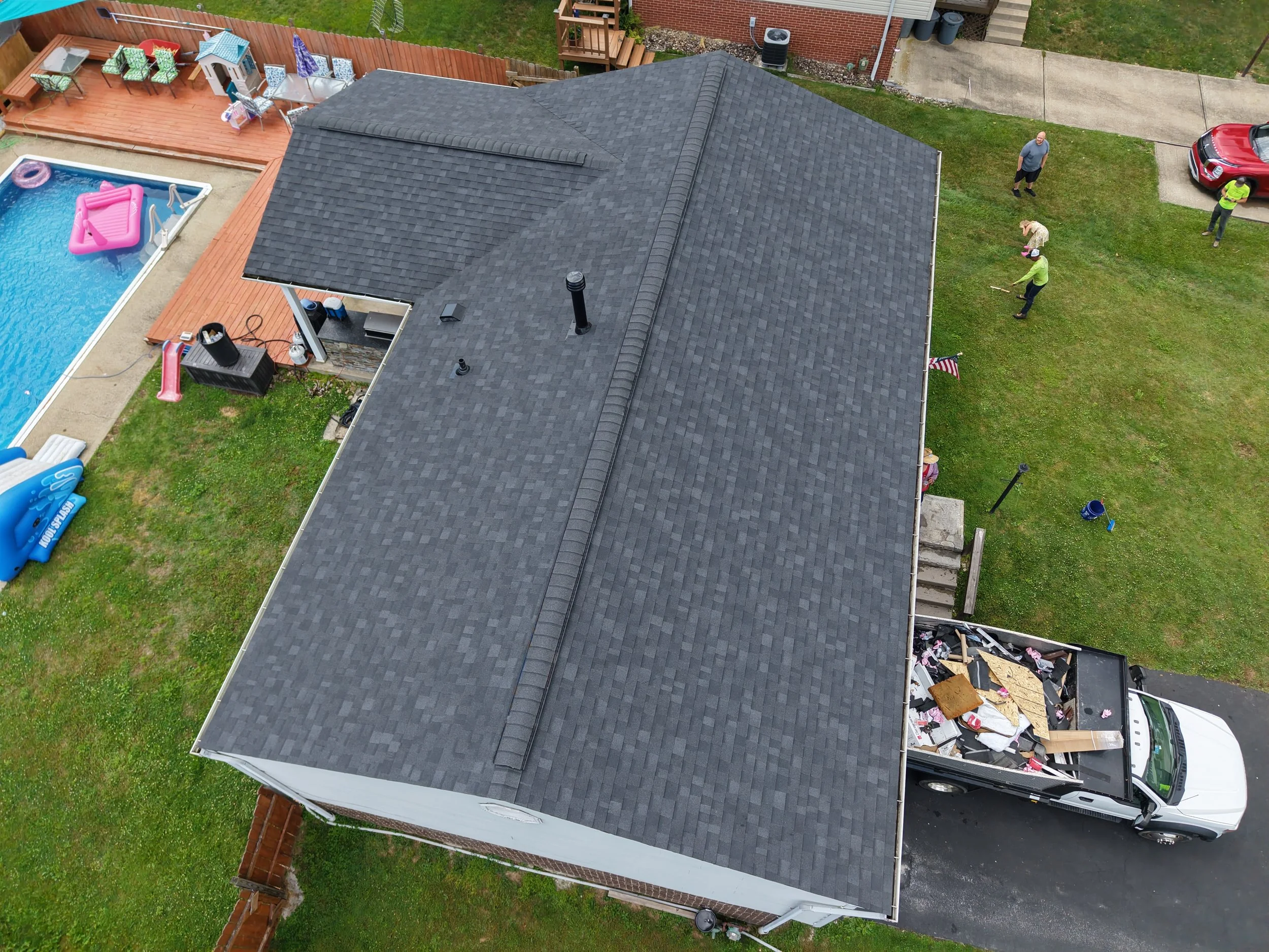 An aerial view of a house with a dark gray shingled roof, a backyard with a swimming pool, a wooden deck with patio furniture, and three people standing and working on the lawn, with some debris and a white pickup truck parked nearby.