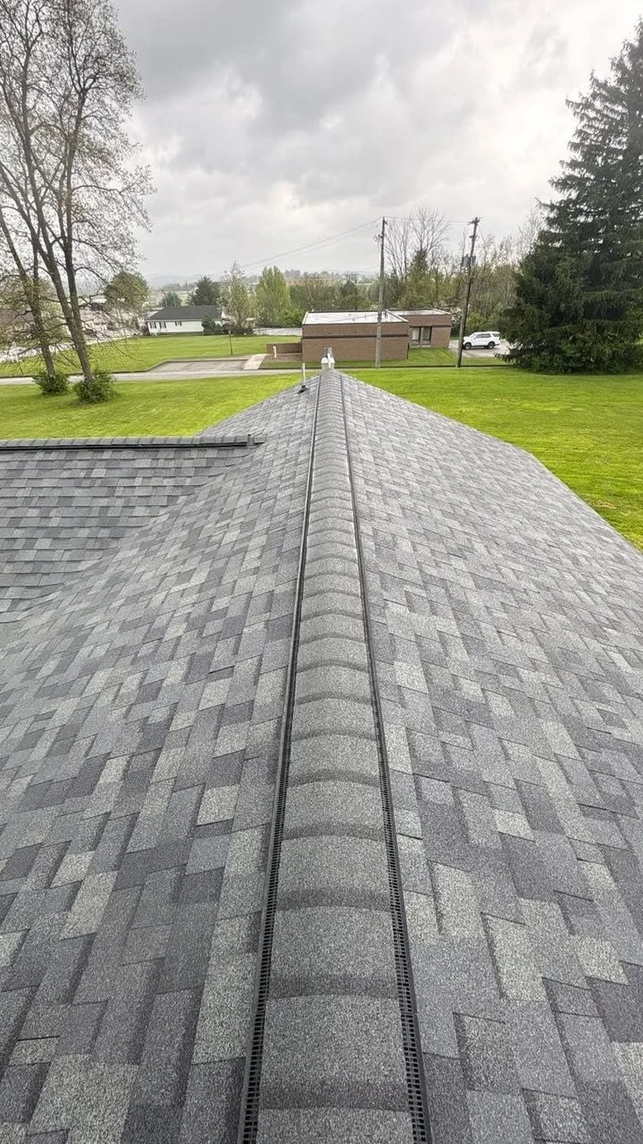 View of a house roof with gray asphalt shingles, central ridge vent, and surrounding grassy yard under a cloudy sky.