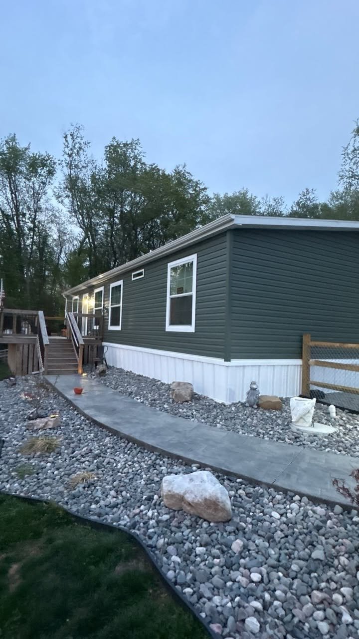 A newly constructed mobile home with a gray exterior and white skirting, featuring multiple windows, a wooden staircase with railings leading to the entrance, and landscaped yard with a concrete pathway, rocks, and decorative items.