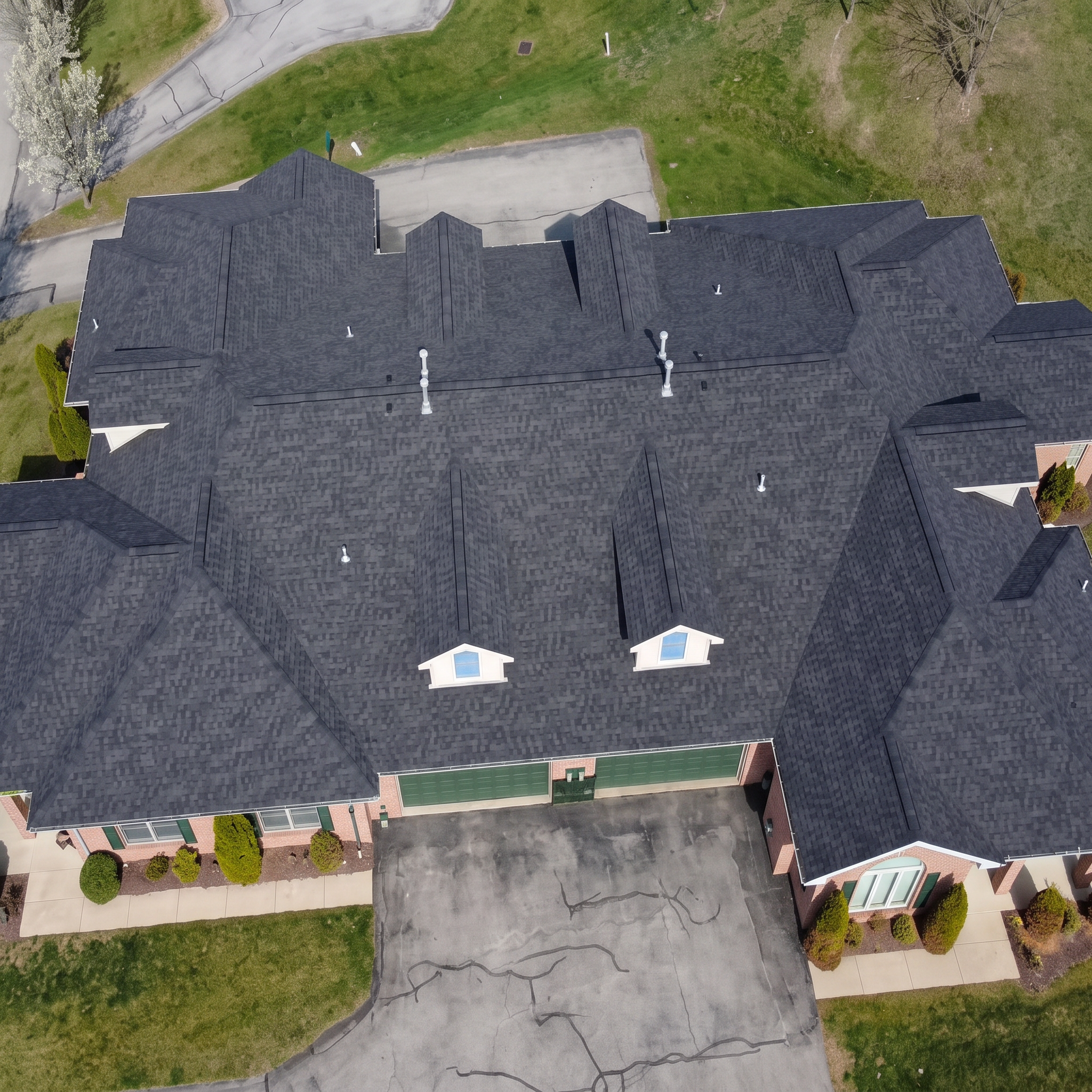 Aerial view of a large residential house with dark shingled roof, multiple chimneys, and windows, surrounded by landscaped yard with green grass, trees, and shrubbery, and a neighboring street.
