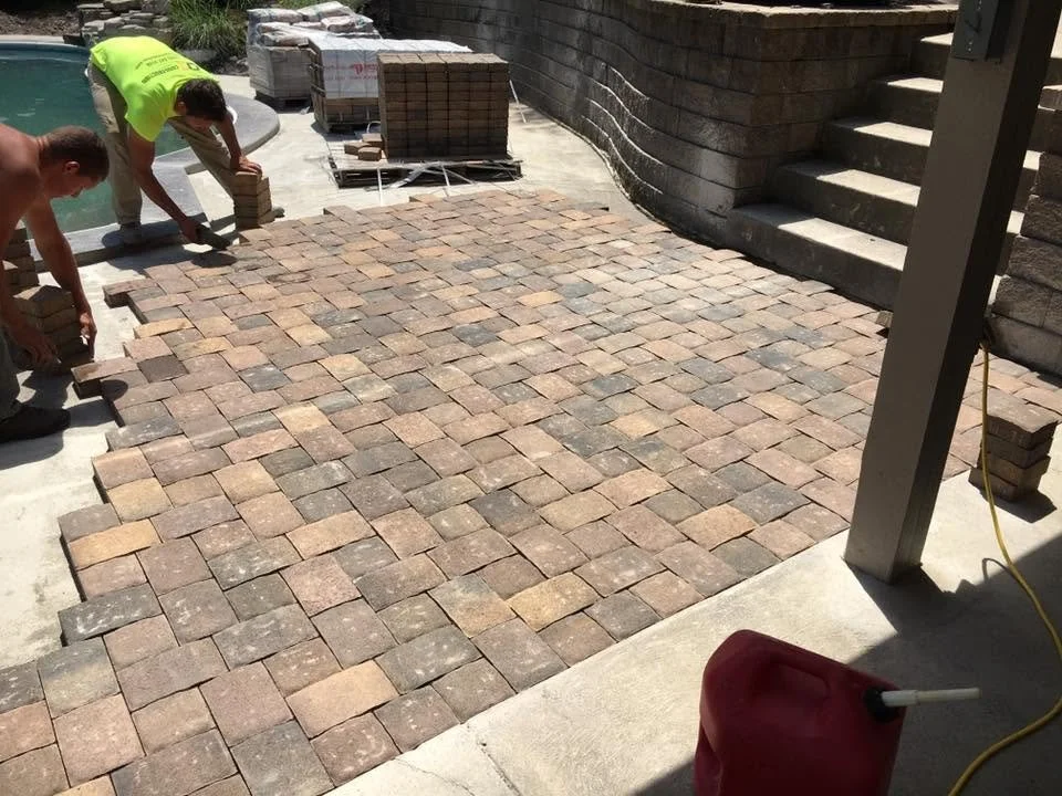Two workers install a brick patio, arranging rectangular pavers in a pattern near a pool.