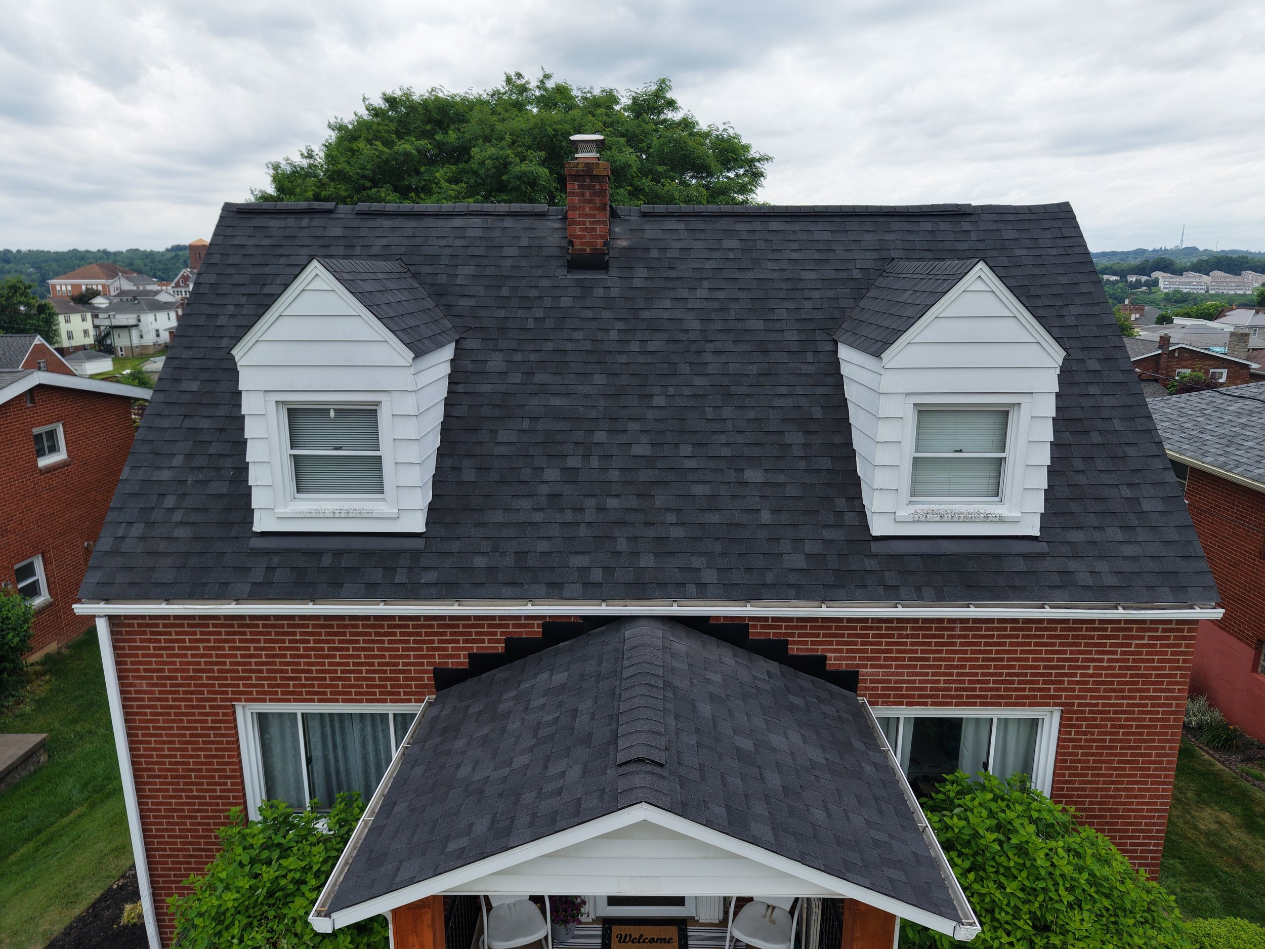 A two-story house with a dark gray gable roof and dormer windows. The house has a brick exterior and white window trims. In the background, there are other houses, trees, and a cloudy sky.