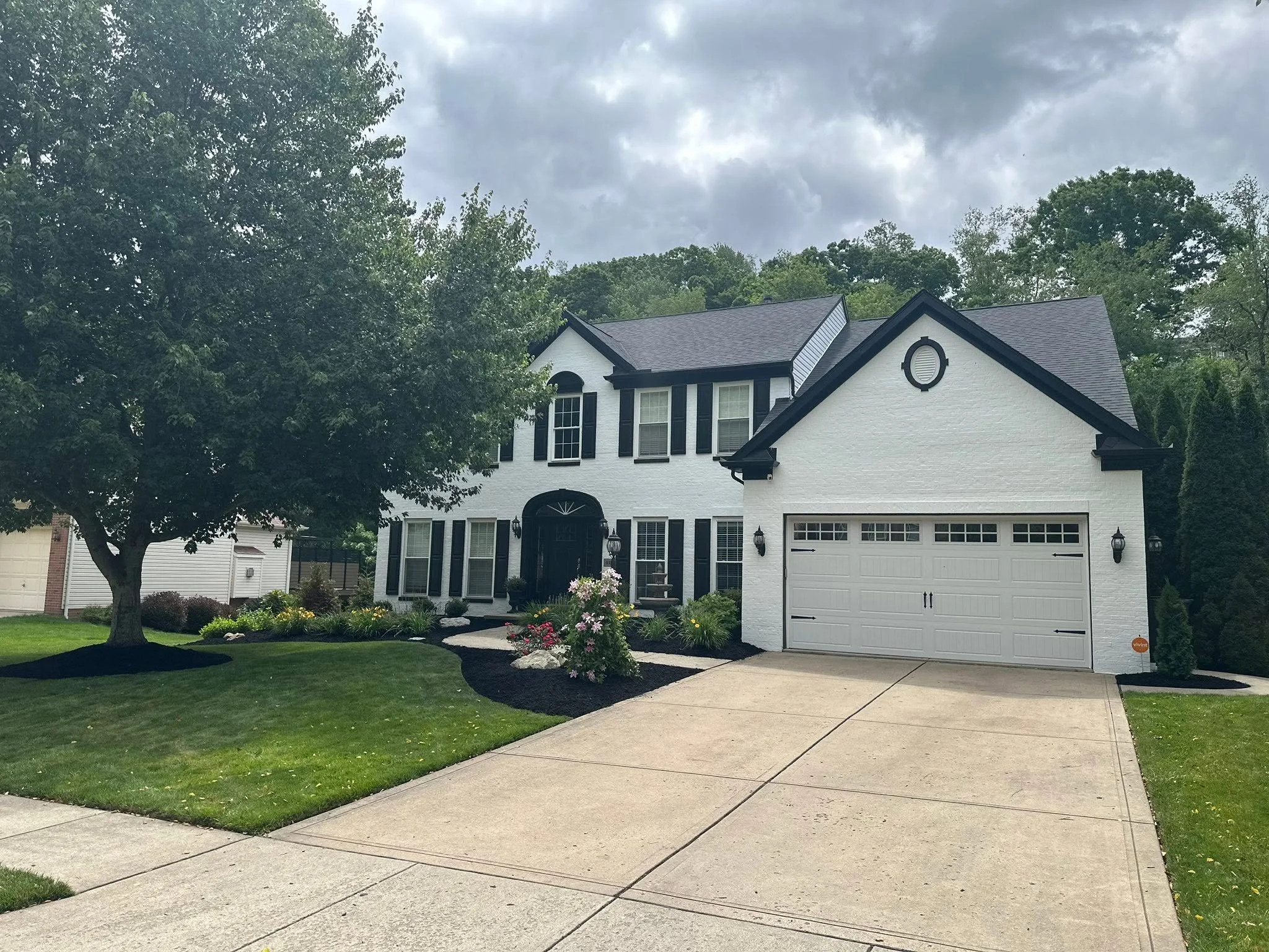 A two-story white house with black shutters and a front garage door. The house is surrounded by green lawn and shrubs, with a large tree on the left and a cloudy sky above.