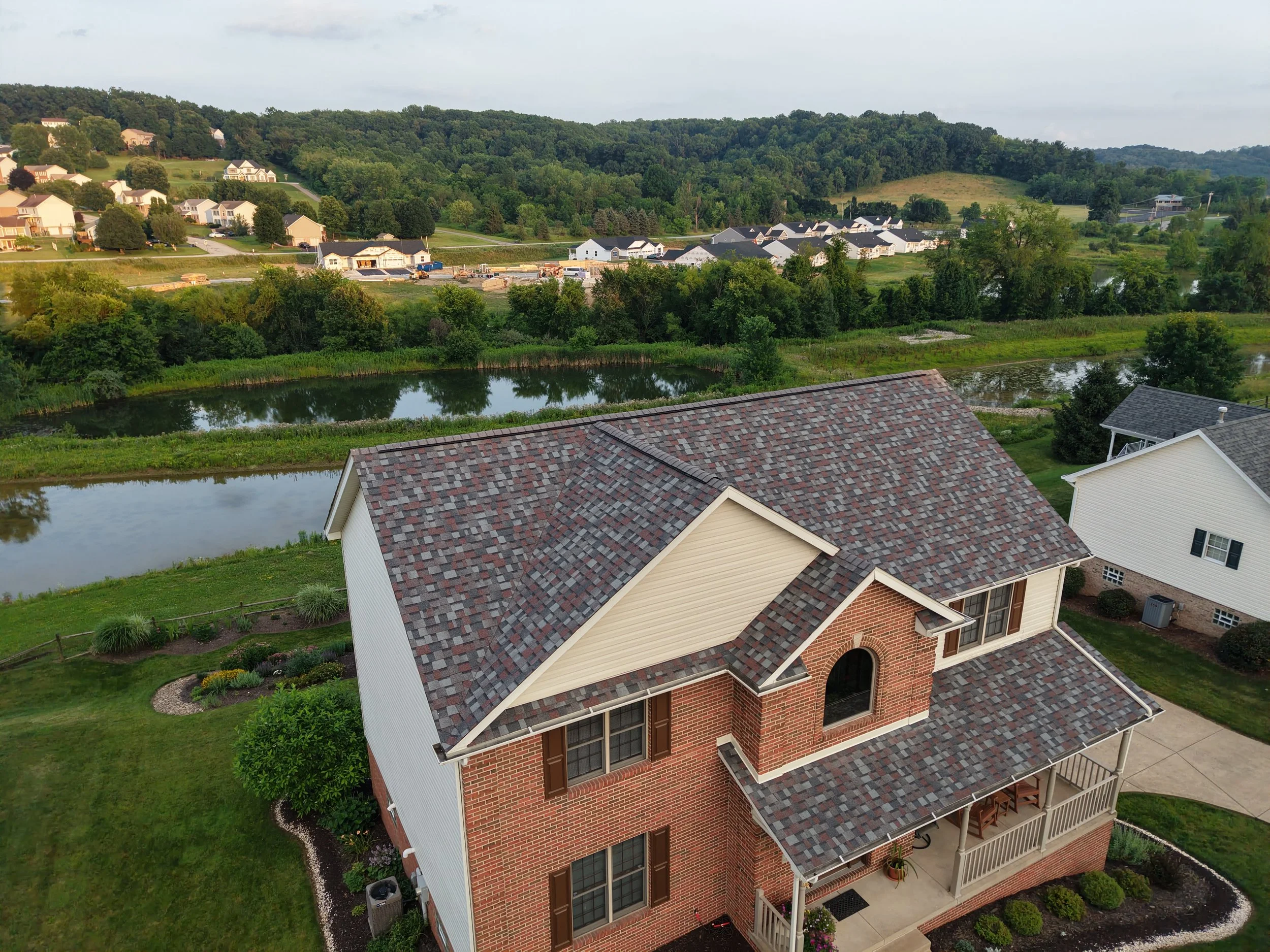 A large two-story house with a brick exterior, white siding, and a multi-gabled roof with shingles, surrounded by a well-maintained lawn and garden. In the background, there is a lake, trees, and a residential neighborhood under a partly cloudy sky.