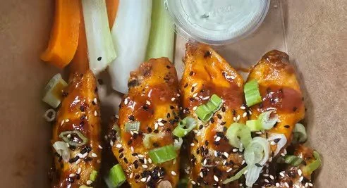 Three buffalo chicken wings topped with sliced green onions and black sesame seeds, along with celery sticks and a small container of dipping sauce.