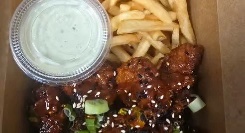 Boneless chicken wings topped with green onions, sesame seeds, and sauce, served with French fries, a side of ranch dipping sauce, in a takeout box.