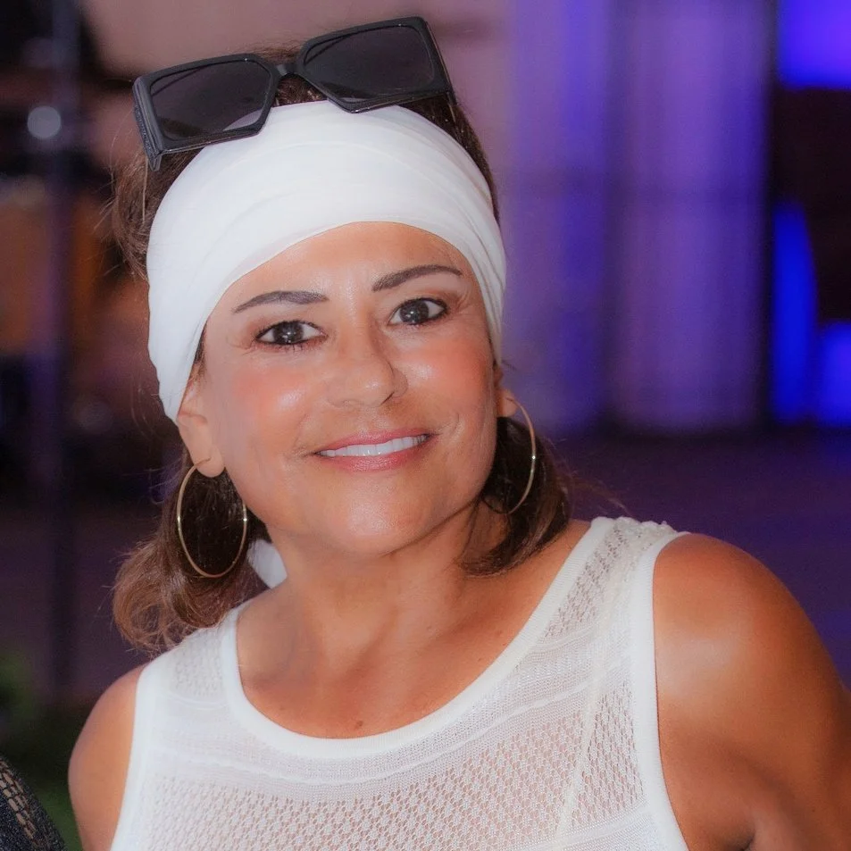 A smiling woman wearing a white headscarf, large hoop earrings, and sunglasses on her head, dressed in a sleeveless white top.
